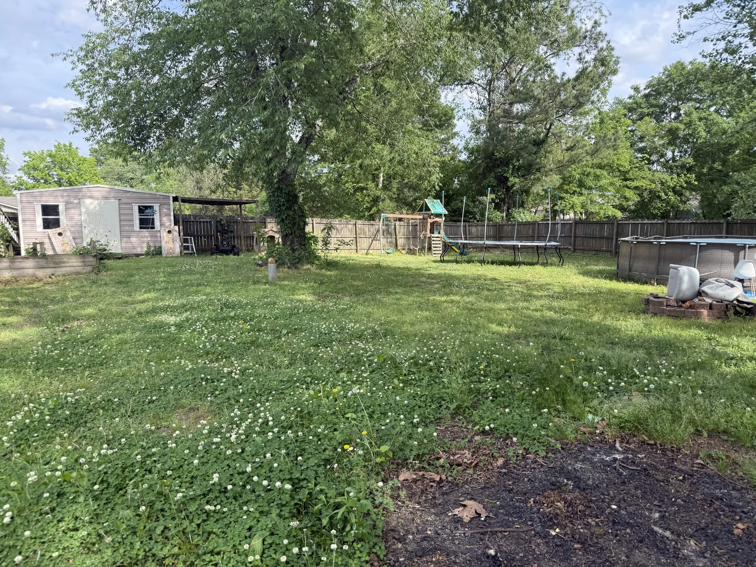 A backyard with a large tree, a small shed, a swing set, a trampoline, and some outdoor furniture on a sunny day.