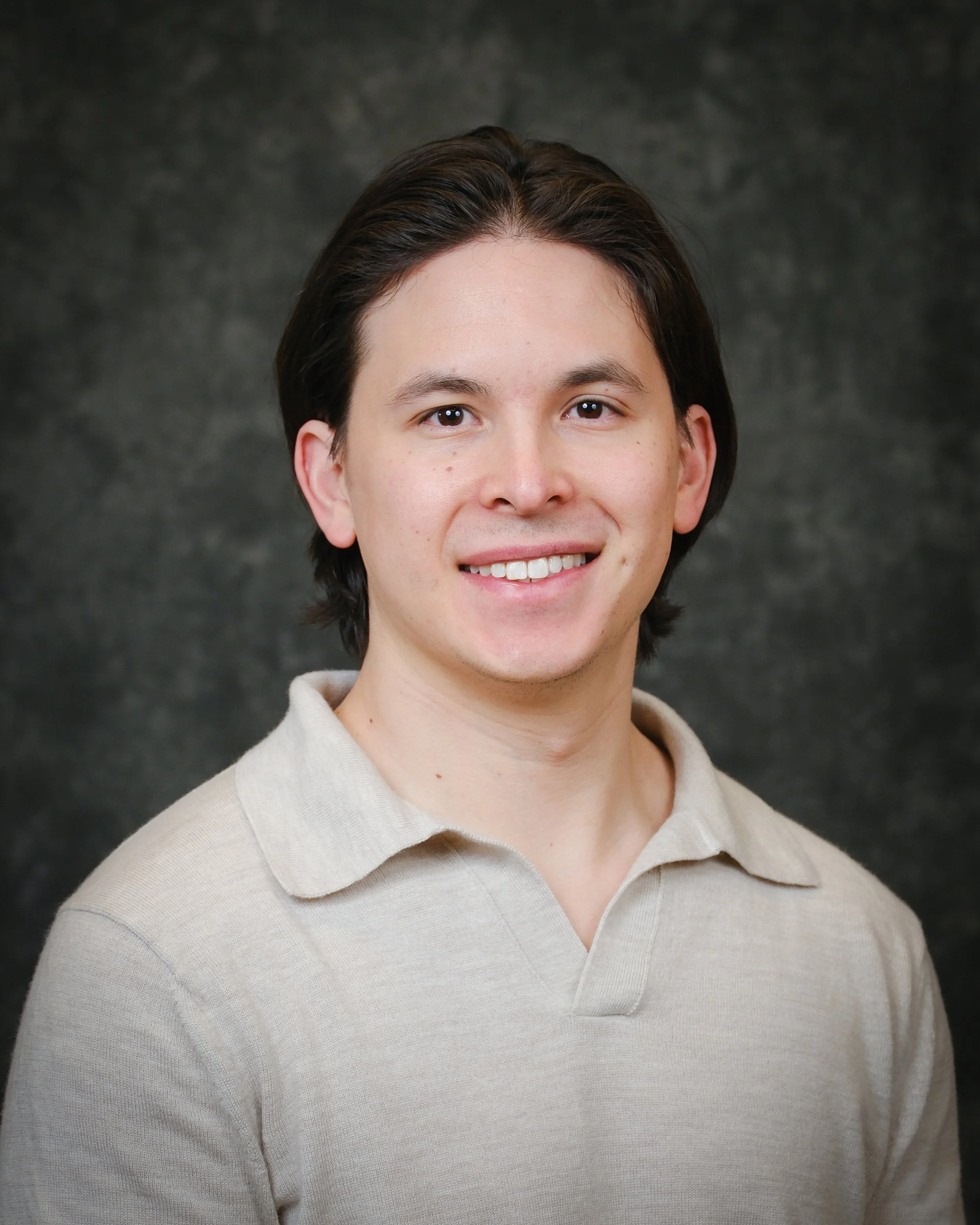 Close-up of a young man with dark brown hair, wearing a beige turtleneck sweater, smiling against a dark grey textured background.