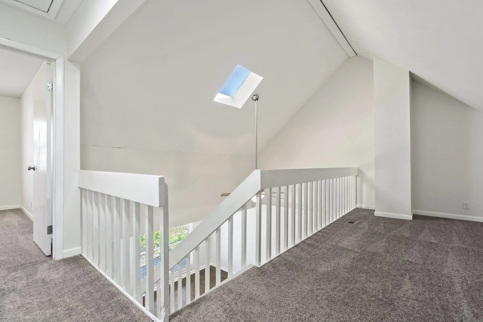 Interior view of a modern upstairs hallway with a skylight, white walls, carpeted floor, white railing, and a door.