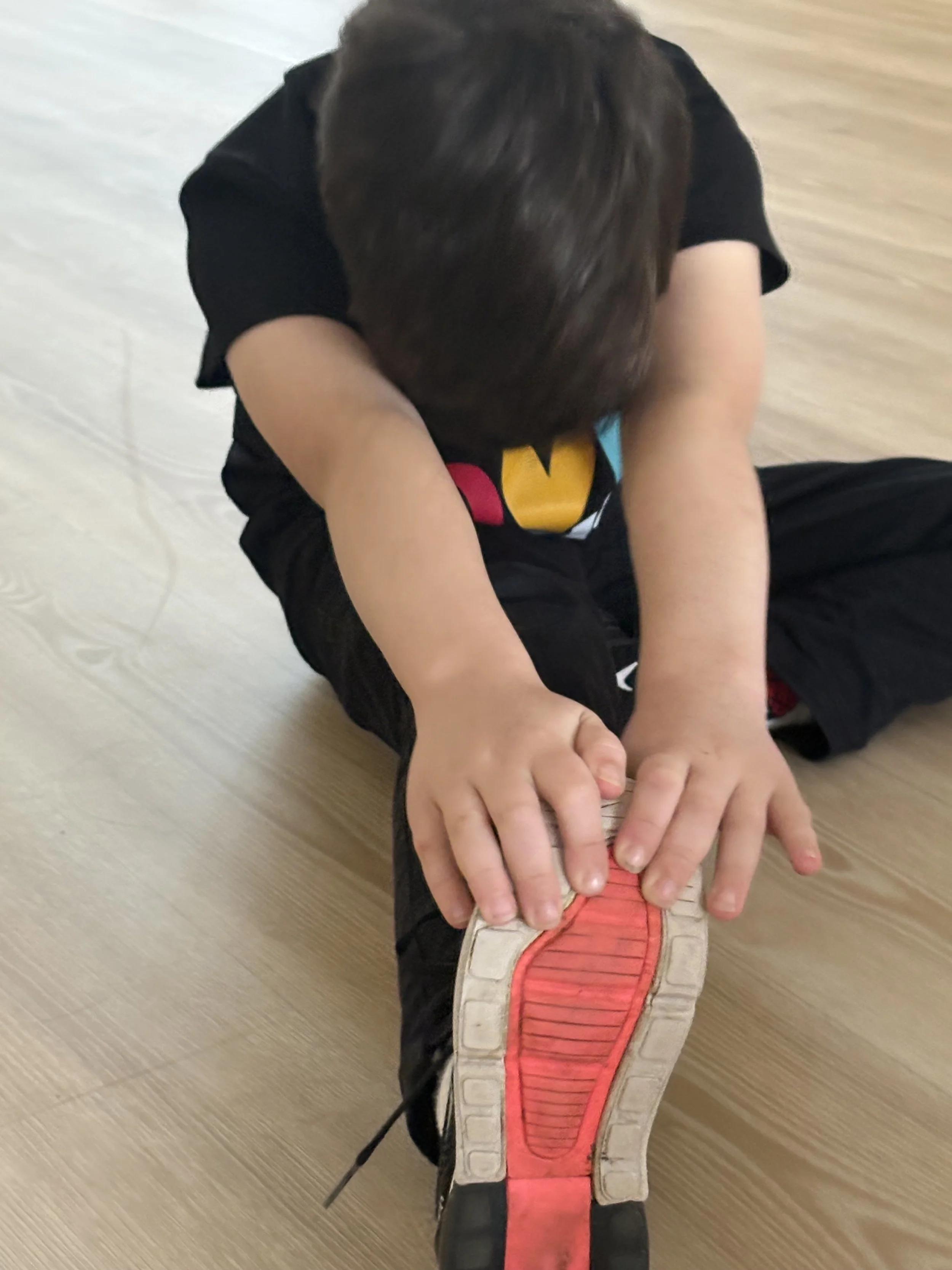 A child sitting on the floor, holding a sneaker, with the sole of the sneaker facing up, showing the red and white design.