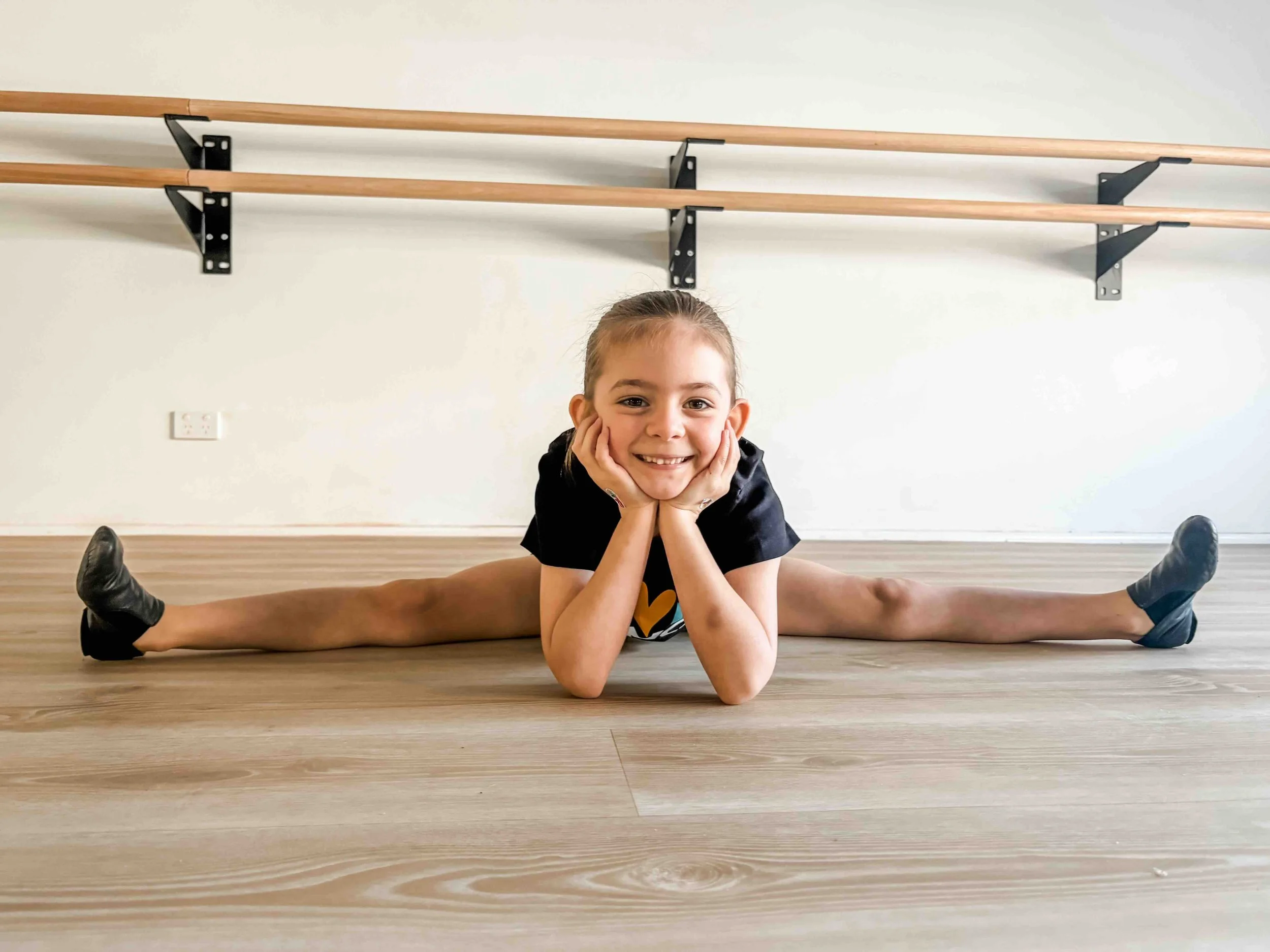 A young girl smiling and lying on her stomach on the floor in a split, supporting her chin with her hands, with wooden ballet bars on the wall behind her.