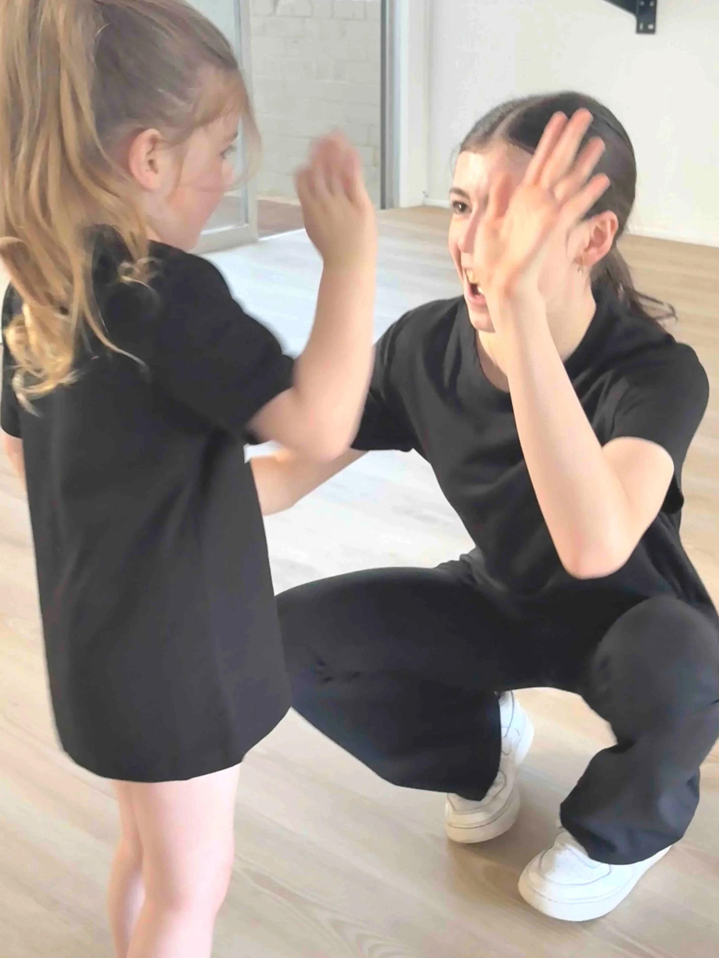 A young girl in black shorts and a black shirt appears to push or playfully hit a woman who is squatting down, dressed in black pants and a black top, with both of them engaging in a playful interaction indoors.