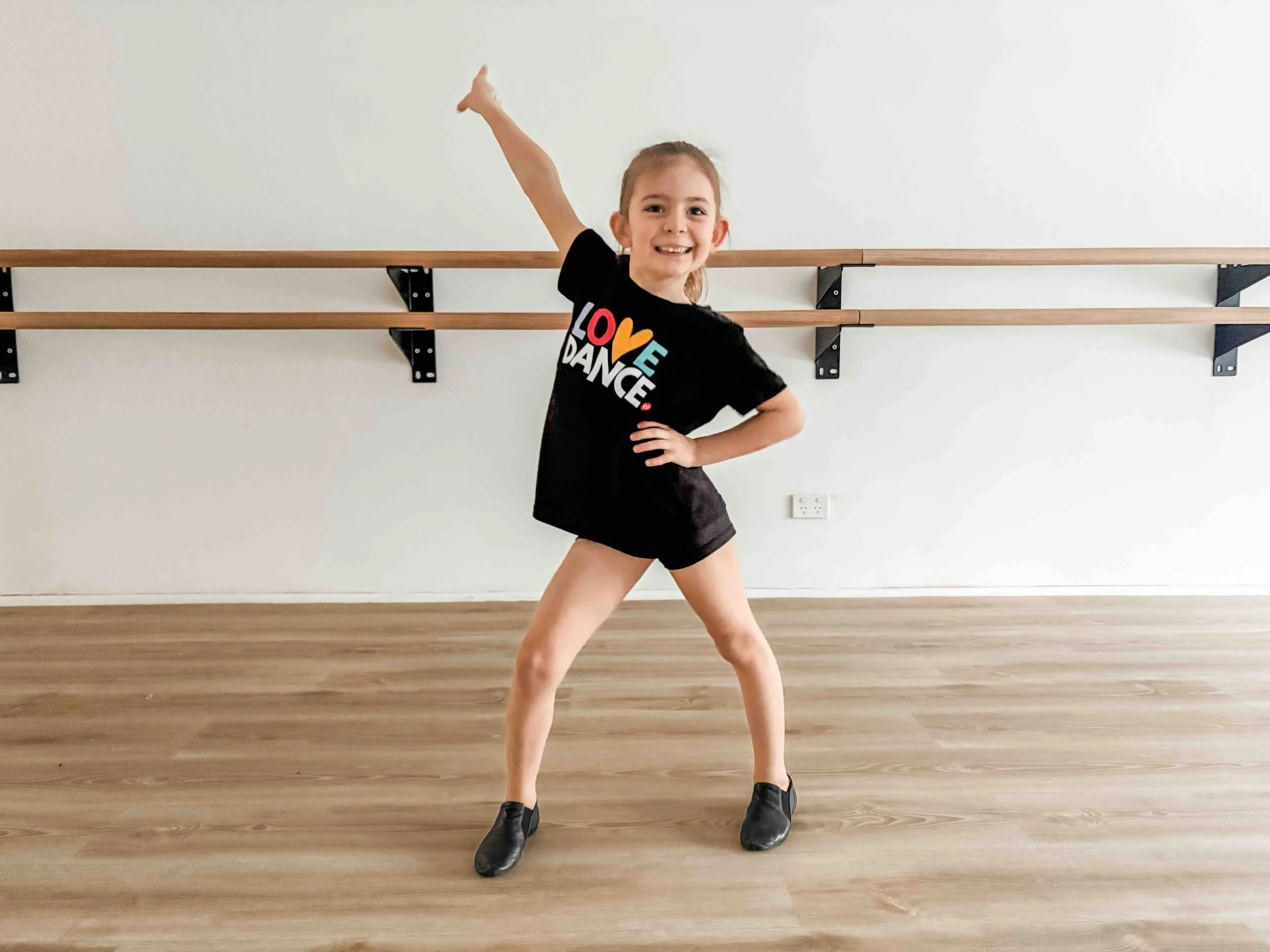 Young girl in a dance studio posing with one hand on her hip and the other raised, wearing a black shirt that says "LOVE DANCE" and black dance shoes, standing in front of a ballet barre against a white wall.