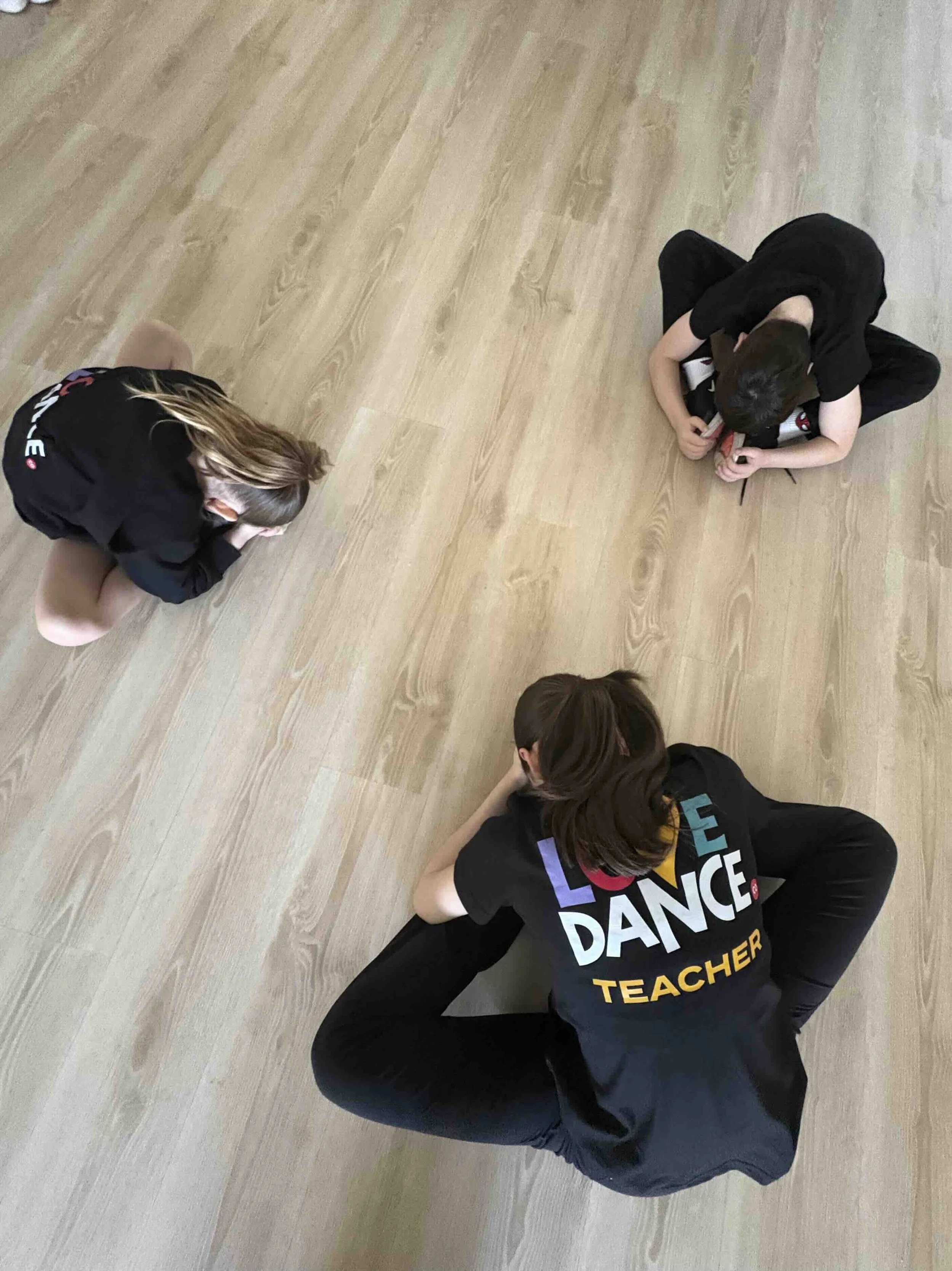 Three children sitting cross-legged on a wooden floor, with one wearing a shirt that says 'LOVE DANCE CO TEACHER'.