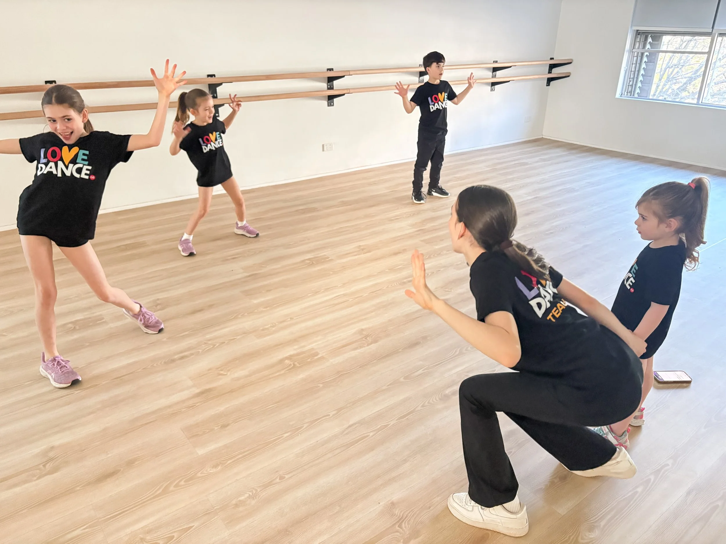 Children and an instructor practicing dance in a dance studio with wooden floors and wall-mounted ballet barres. The children are dressed in black shirts reading 'LOVE DANCE' and are engaging in a warm-up or dance routine.
