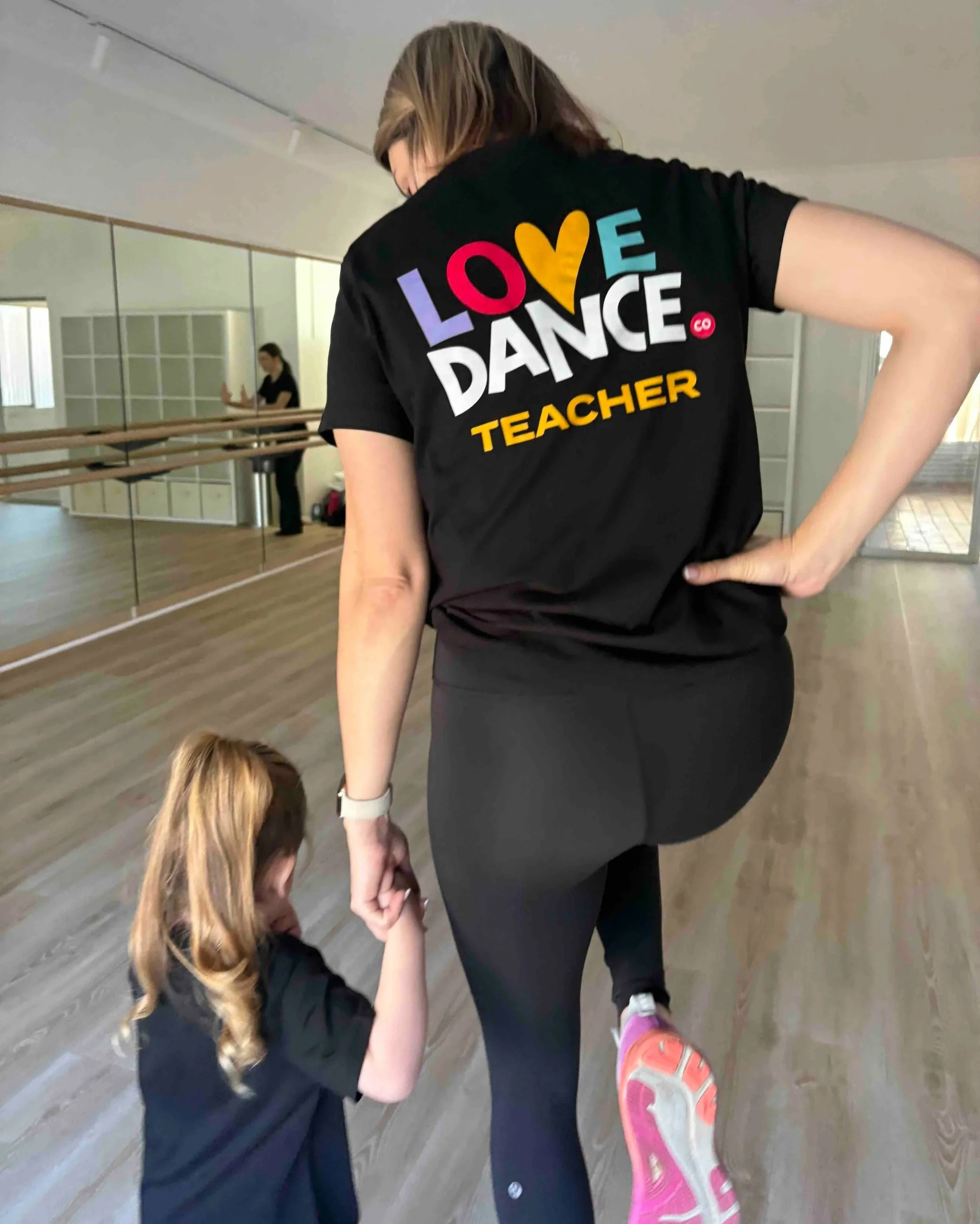 Dance instructor in black T-shirt labeled 'Love Dance Teacher' assists young girl with brown hair and black jacket, wearing pink and white sneakers, in a dance studio with mirrors and wooden floor.