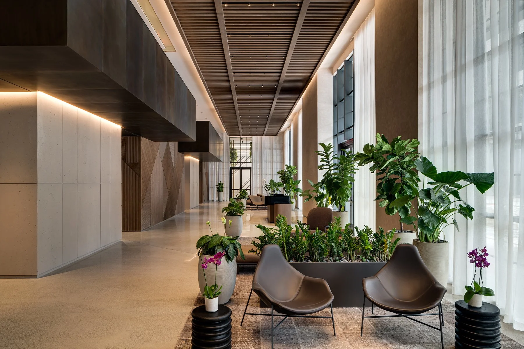 Interior view of a modern lobby or waiting area with large windows, white curtains, and numerous potted green plants. Contains stylish chairs and a wooden ceiling with integrated lighting.