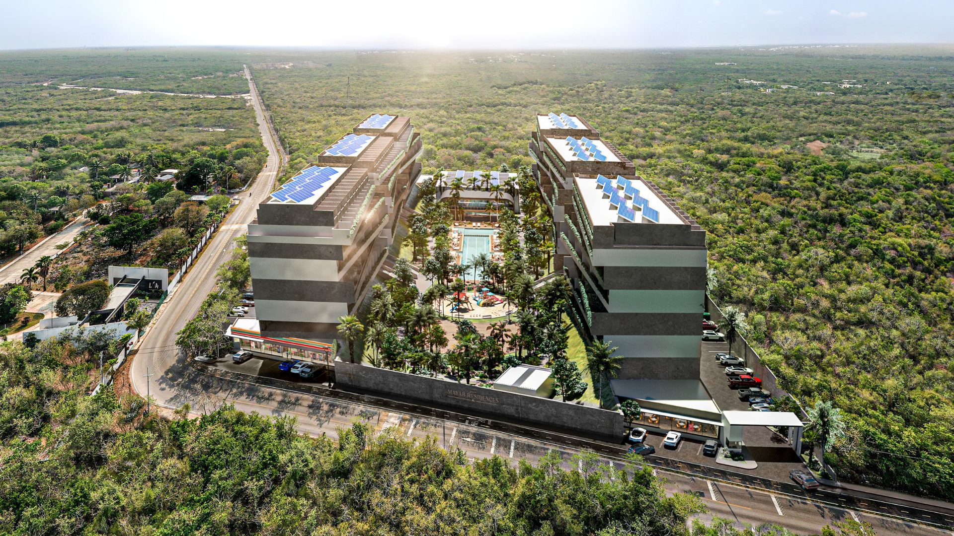 Vista aérea de un complejo residencial con edificios altos, piscina central, áreas verdes y parques infantiles, rodeado de áreas boscosas y caminos asfaltados.