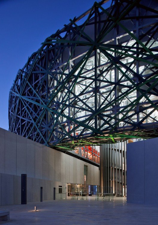 Estructura moderna de metal con formas geométricas, iluminada desde dentro, en un entorno urbano durante el atardecer.