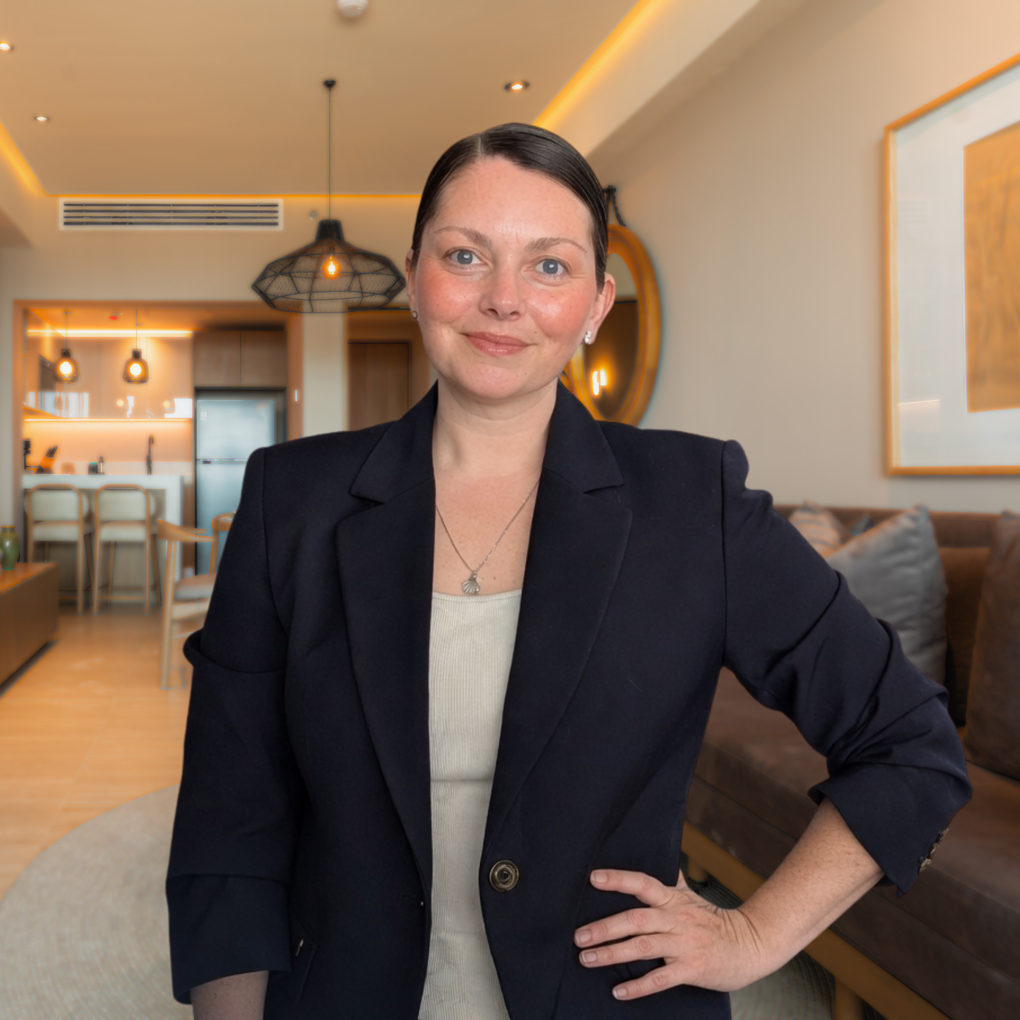 A woman with short dark hair wearing a black blazer and a necklace with a pendant standing in a modern interior with warm lighting, a round mirror, wooden furniture, and pendant lights
