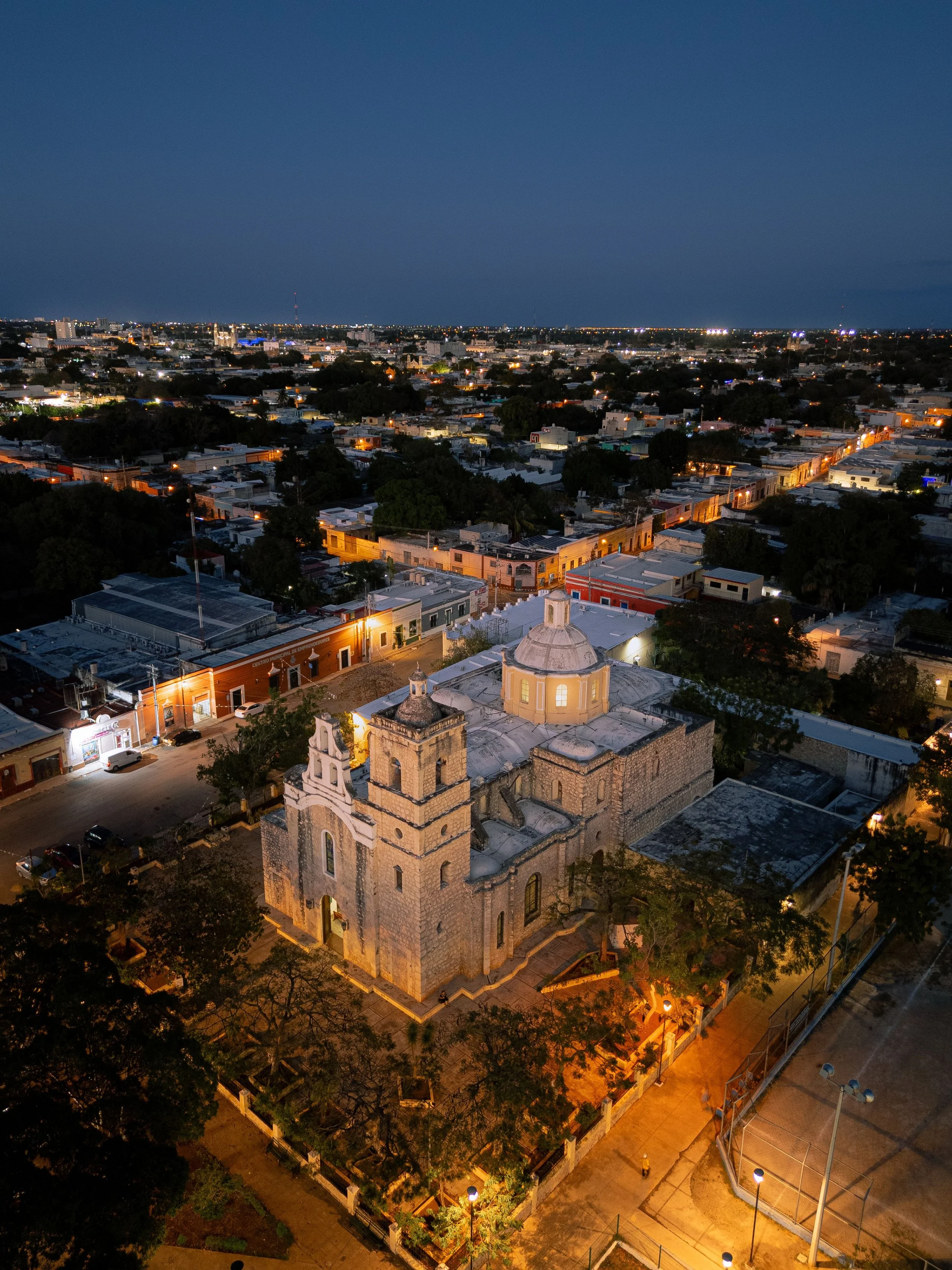 Vista nocturna de una ciudad con calles iluminadas y un cielo despejado, donde destaca una iglesia antigua en primer plano y edificios modernos en el fondo.