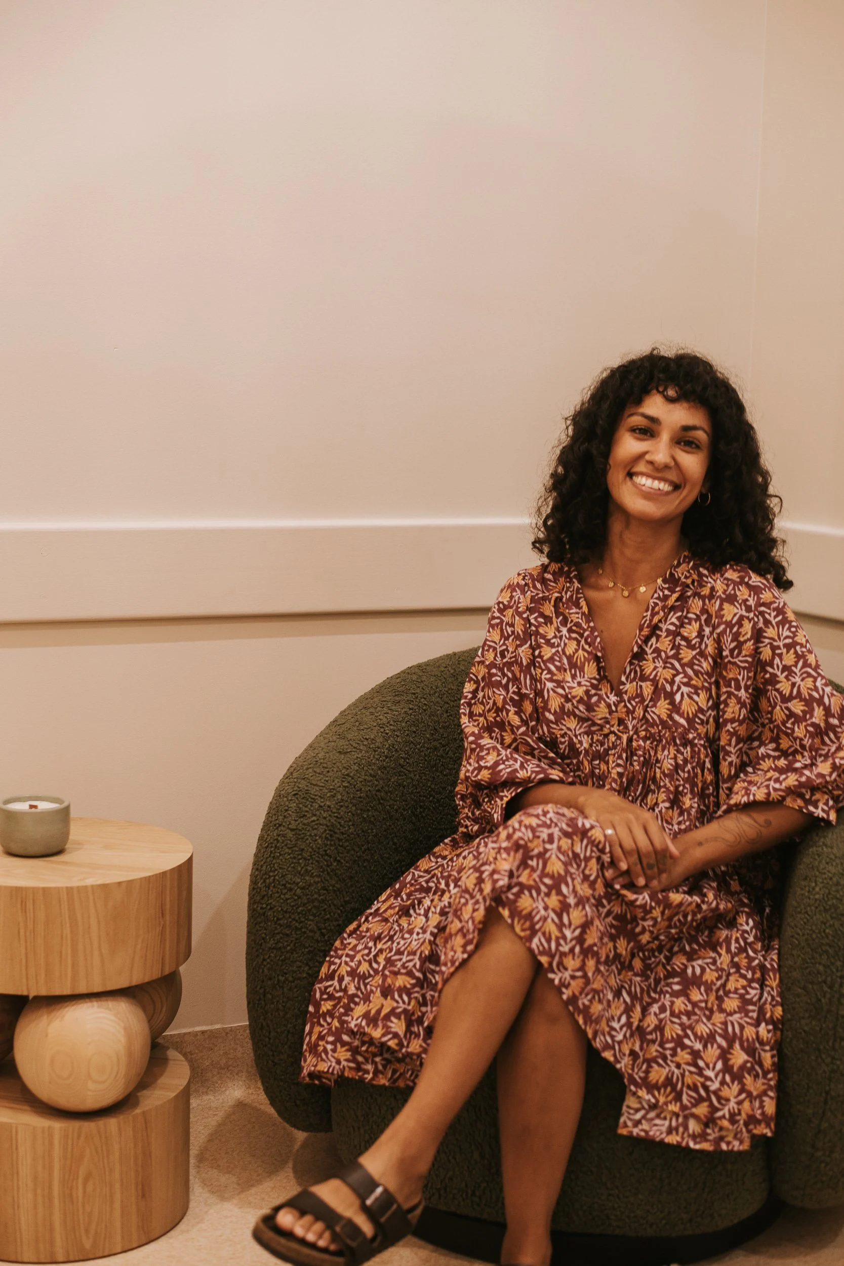 A woman with curly hair smiling, sitting on a dark green textured armchair, wearing a matching patterned dress with yellow and maroon leaf motifs, black sandals, and jewelry, in a cozy room with beige walls and a wooden side table with a small candle.