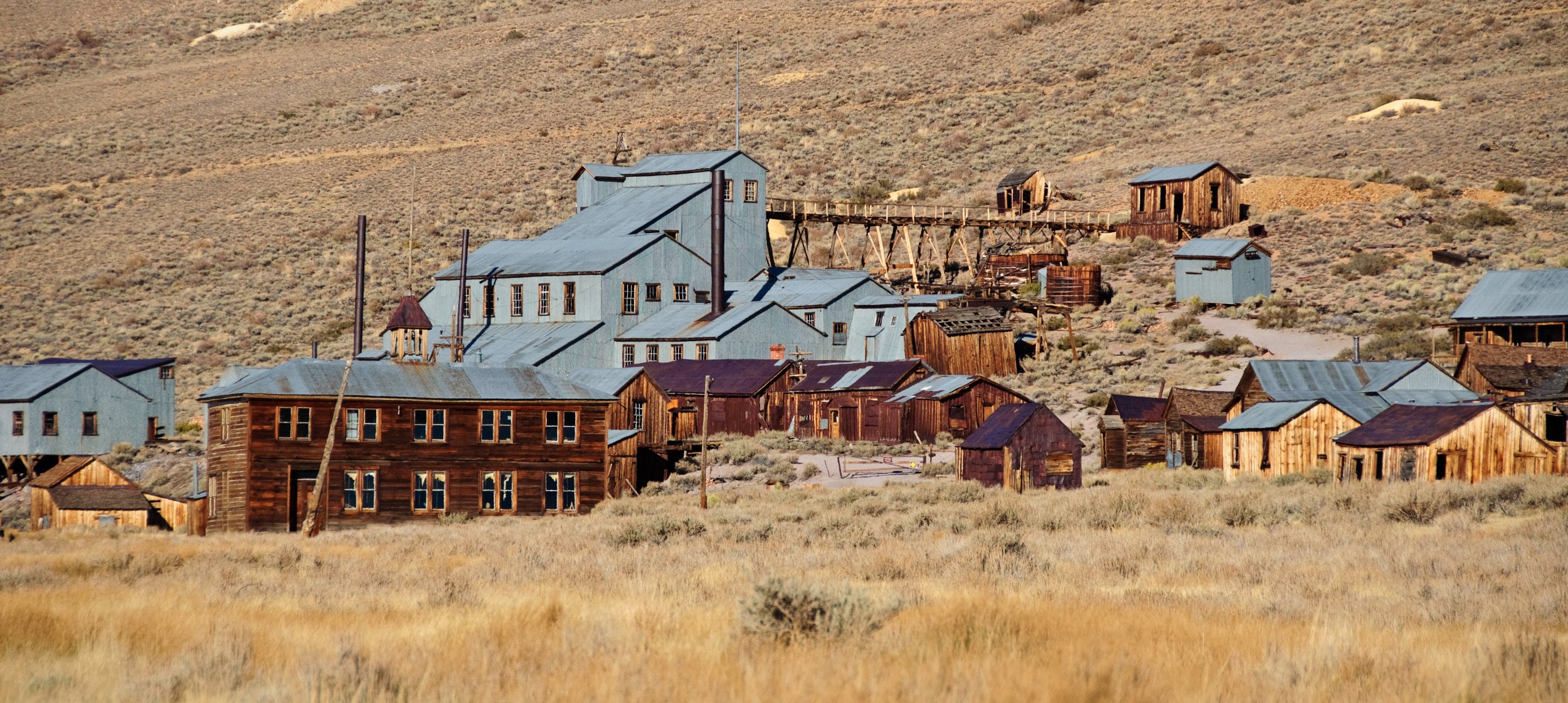 An old ghost town with wooden buildings and a large blue industrial structure in a desert landscape with hills in the background.