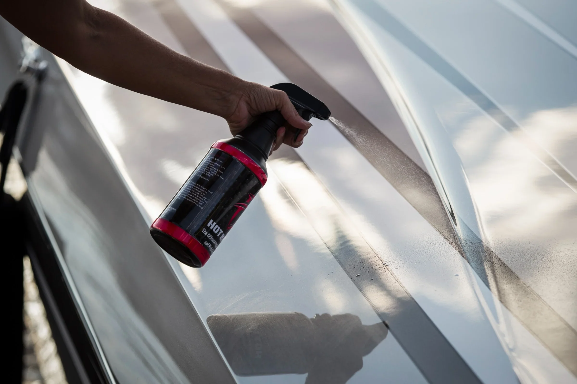 Close-up of a hand holding a spray bottle while applying a cleaning product to the surface of a car.