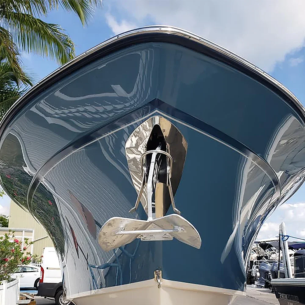 Front view of a large blue boat with a shiny metallic anchor hanging from the bow, set against a background of blue sky, palm trees, and other boats.