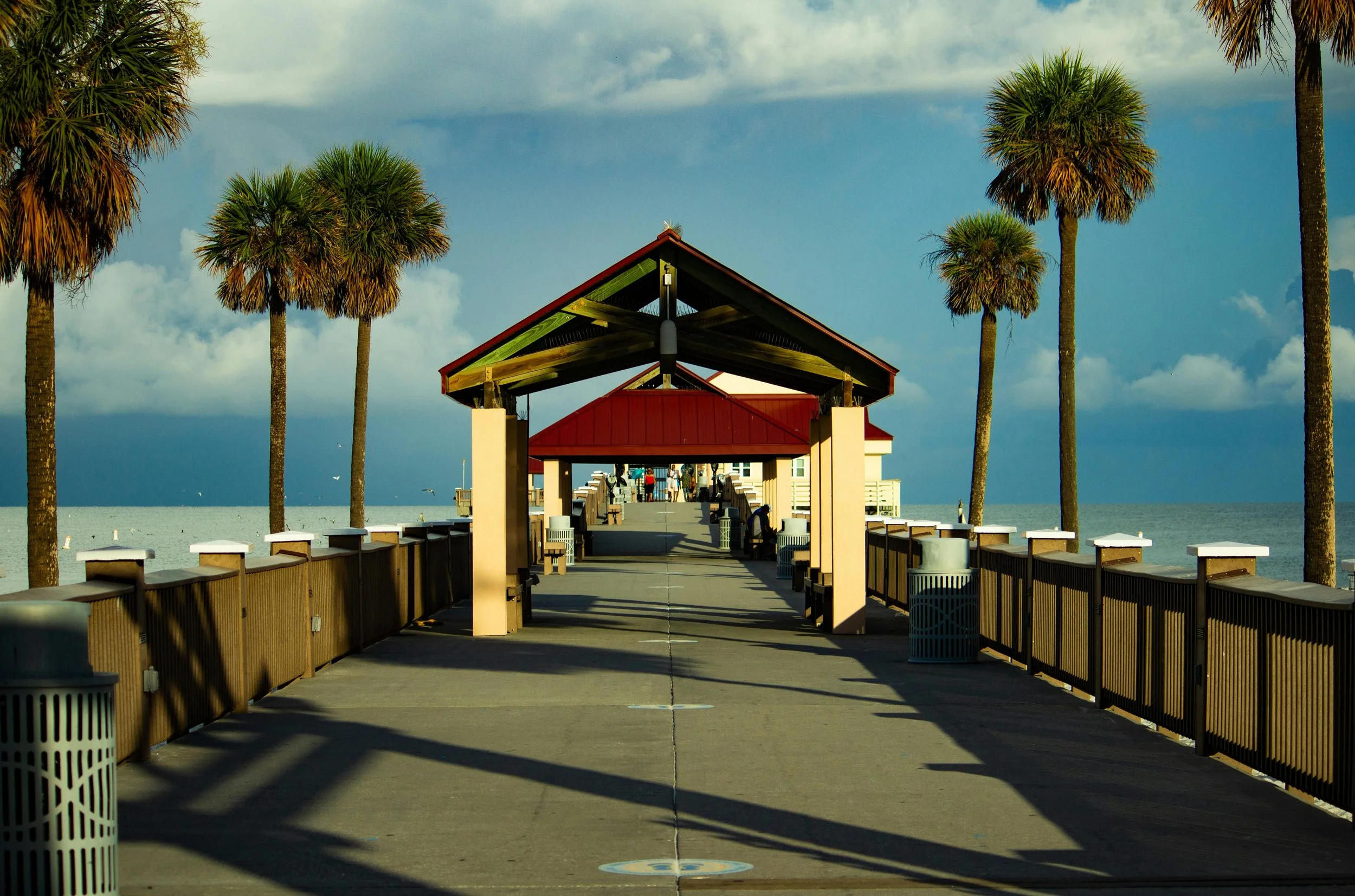 A boardwalk leading to a pier with a pavilion, surrounded by palm trees, on a cloudy day near the ocean.