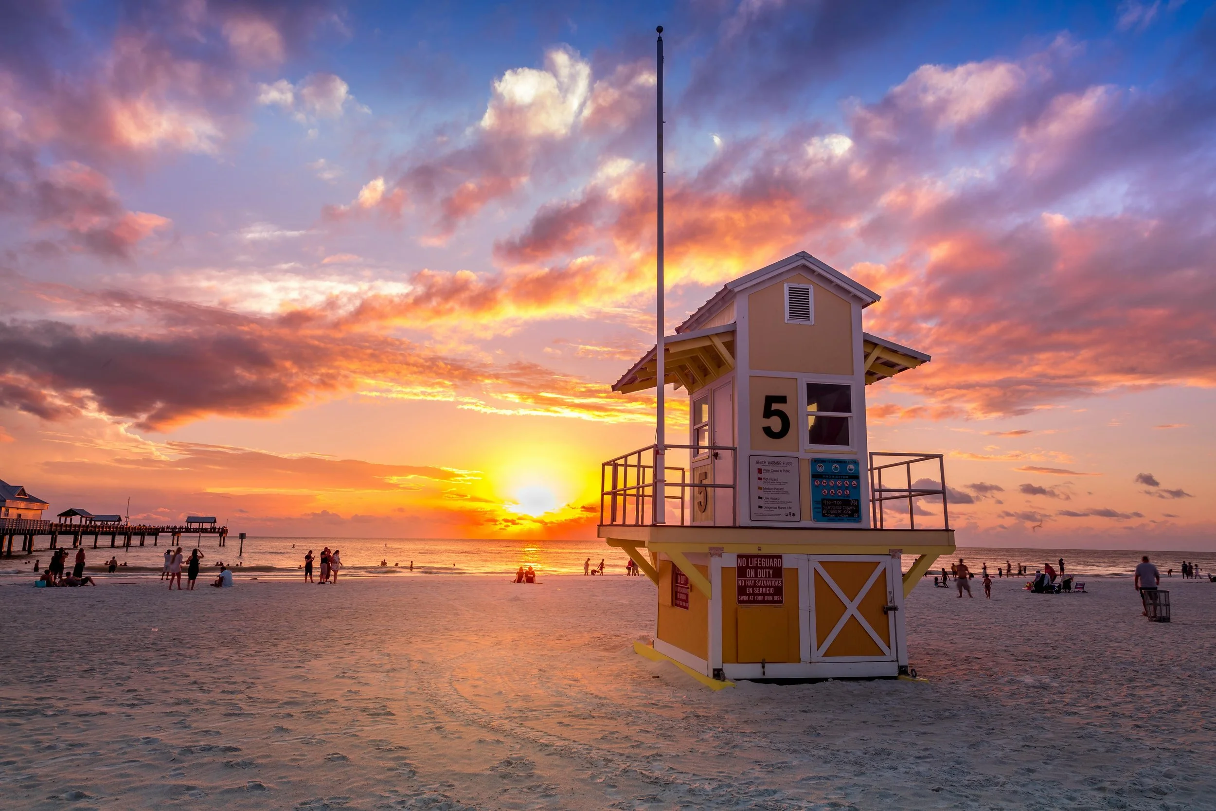 A lifeguard tower with the number 5 on it, set against a sunset sky with colorful clouds at the beach. There are people walking and sitting on the sand, with a pier visible in the background.