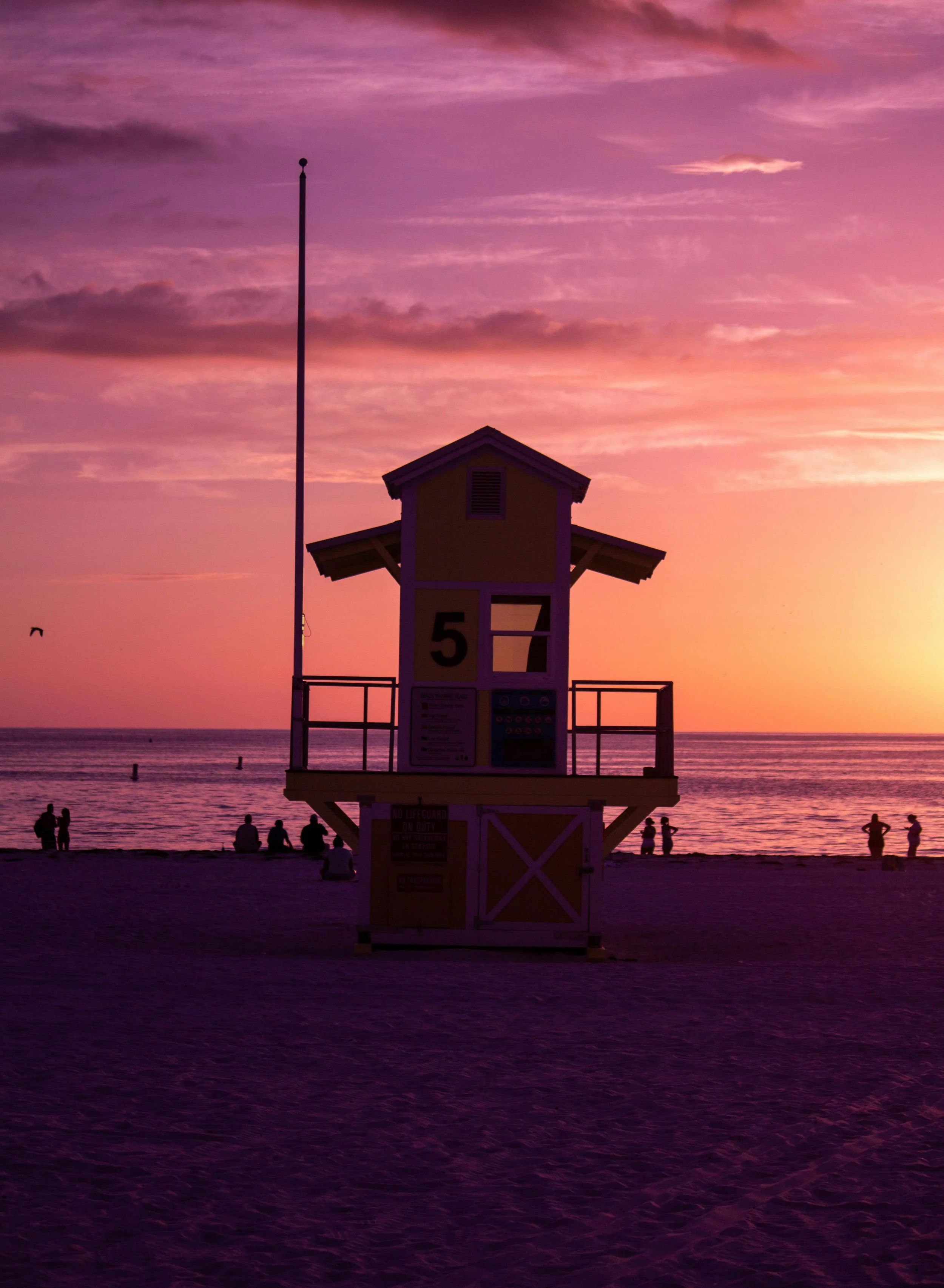 A colorful sunset on the beach with a yellow lifeguard tower marked with the number 57 in the foreground, people sitting and standing along the shoreline, and a few birds flying in the sky.