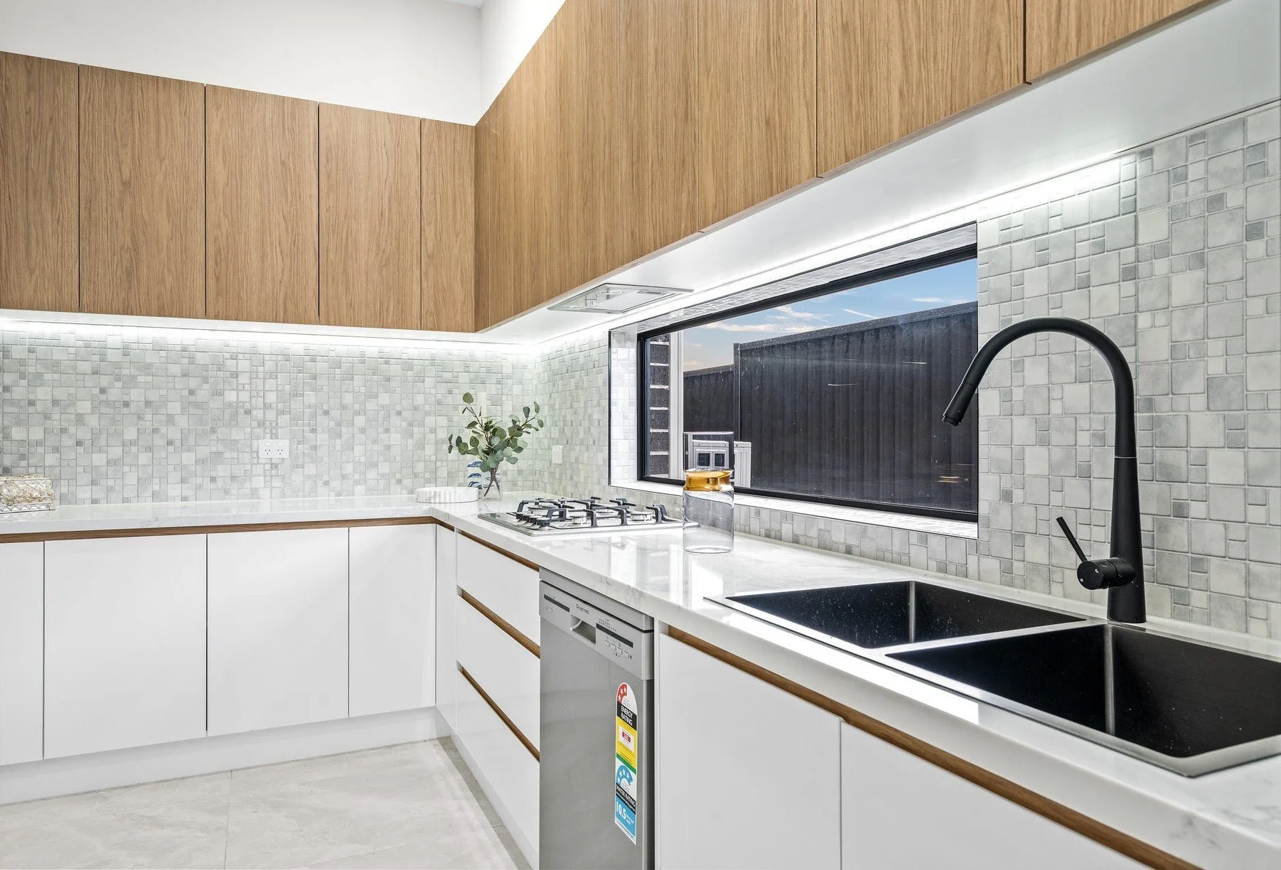 Modern kitchen with white cabinets, black sink, black faucet, marble countertop, mosaic tile backsplash, wooden upper cabinets, and a large window showing an outdoor space.