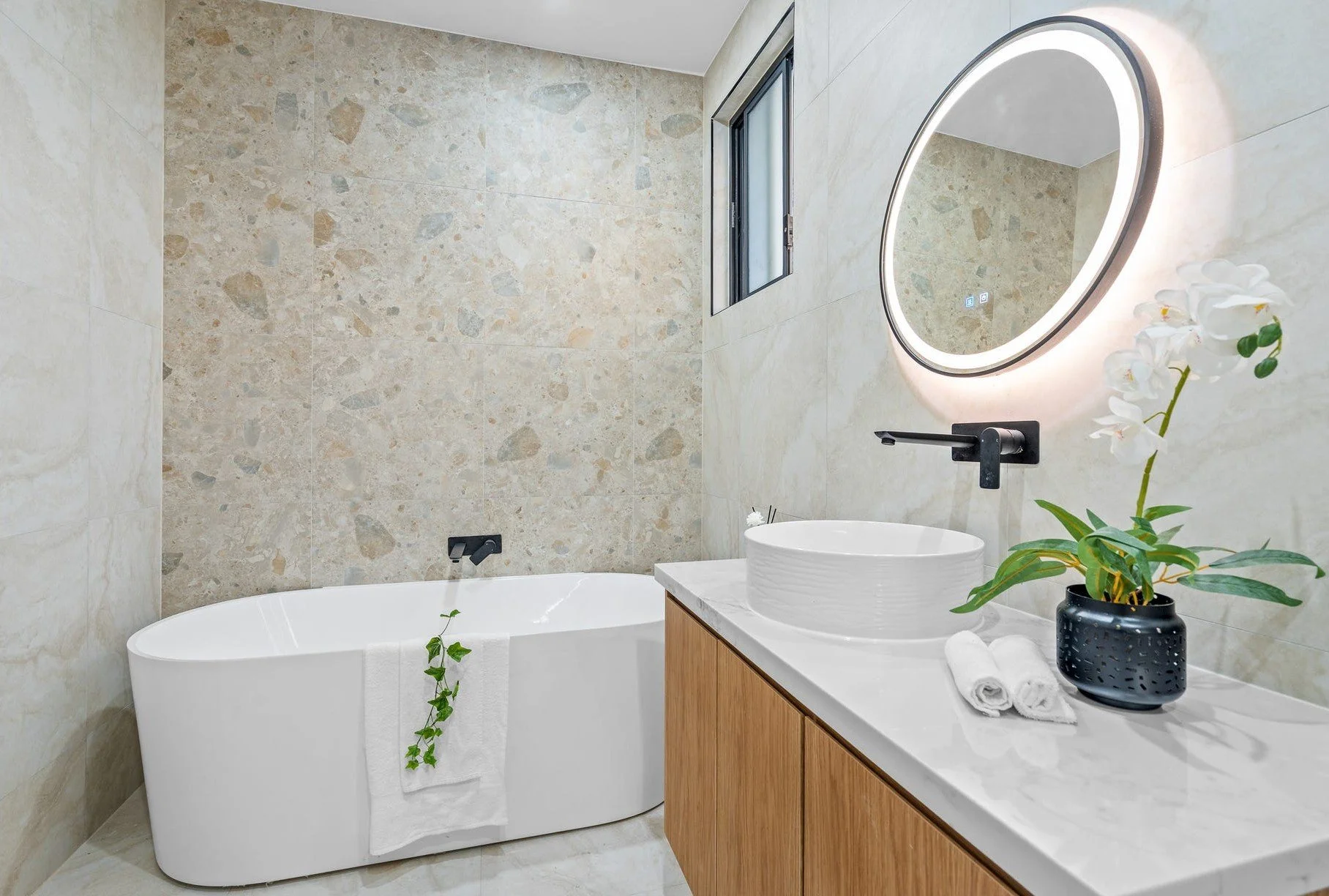 Modern bathroom with a freestanding bathtub, a wooden vanity with a vessel sink, a backlit round mirror, a potted orchid, and a small window, with beige stone tiles on the walls.