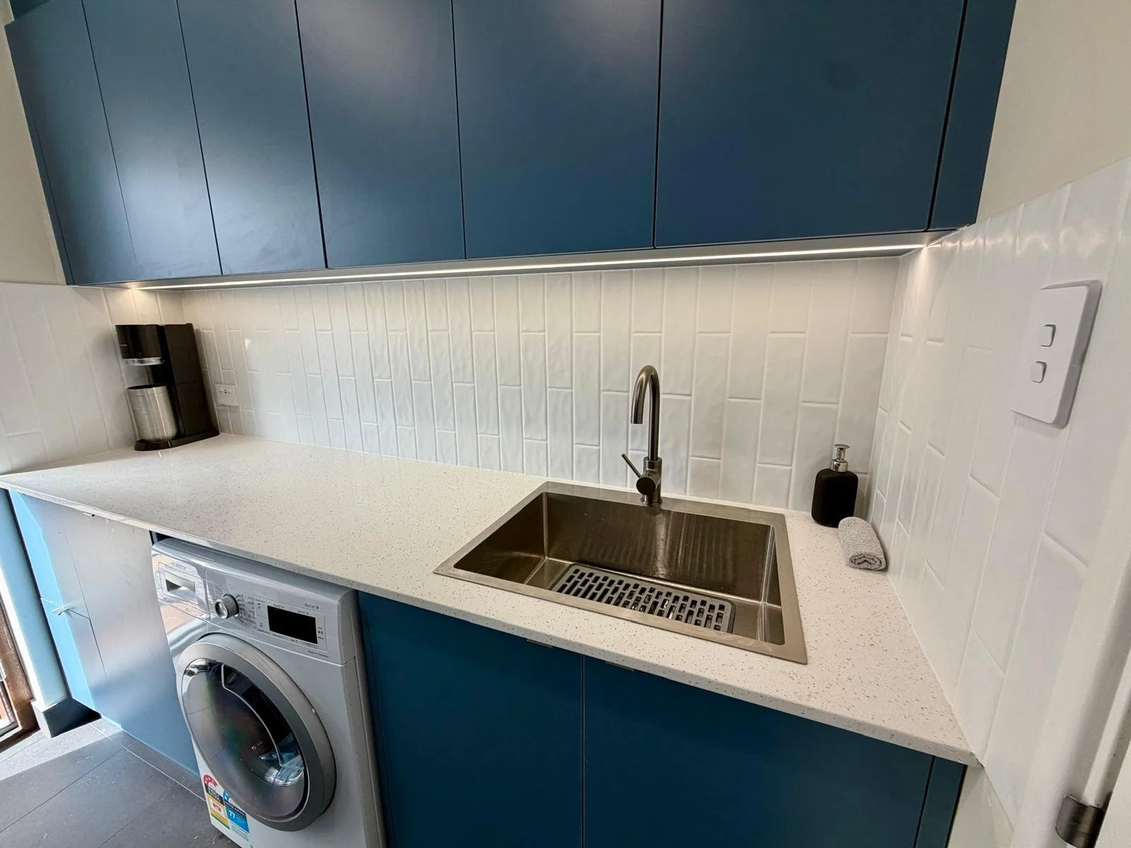 Modern kitchen with blue cabinets, white countertop, stainless steel sink, black soap dispenser, gray towel, washing machine, coffee machine, white tiled backsplash, and electrical outlet.