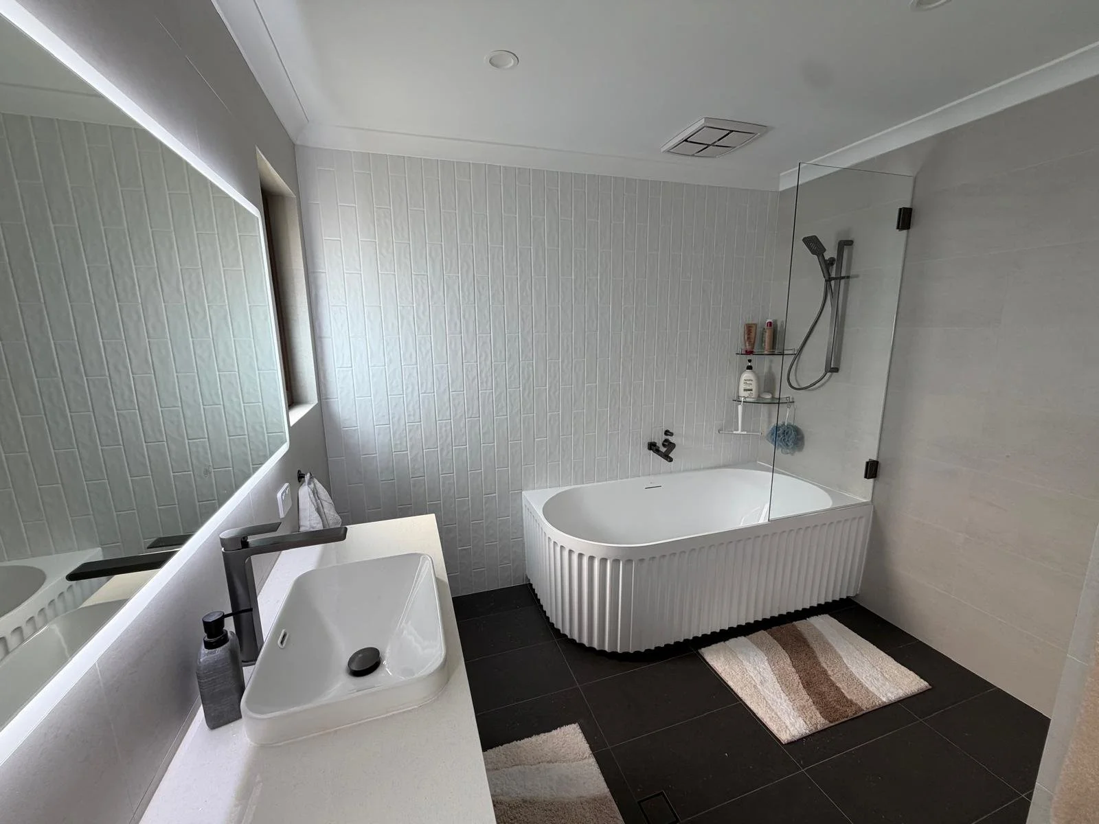 Modern bathroom with a white bathtub, glass shower enclosure, black fixtures, white sink with black faucet, large mirror, white tiled walls, and a brown and white striped bath mat on dark tiled floor.