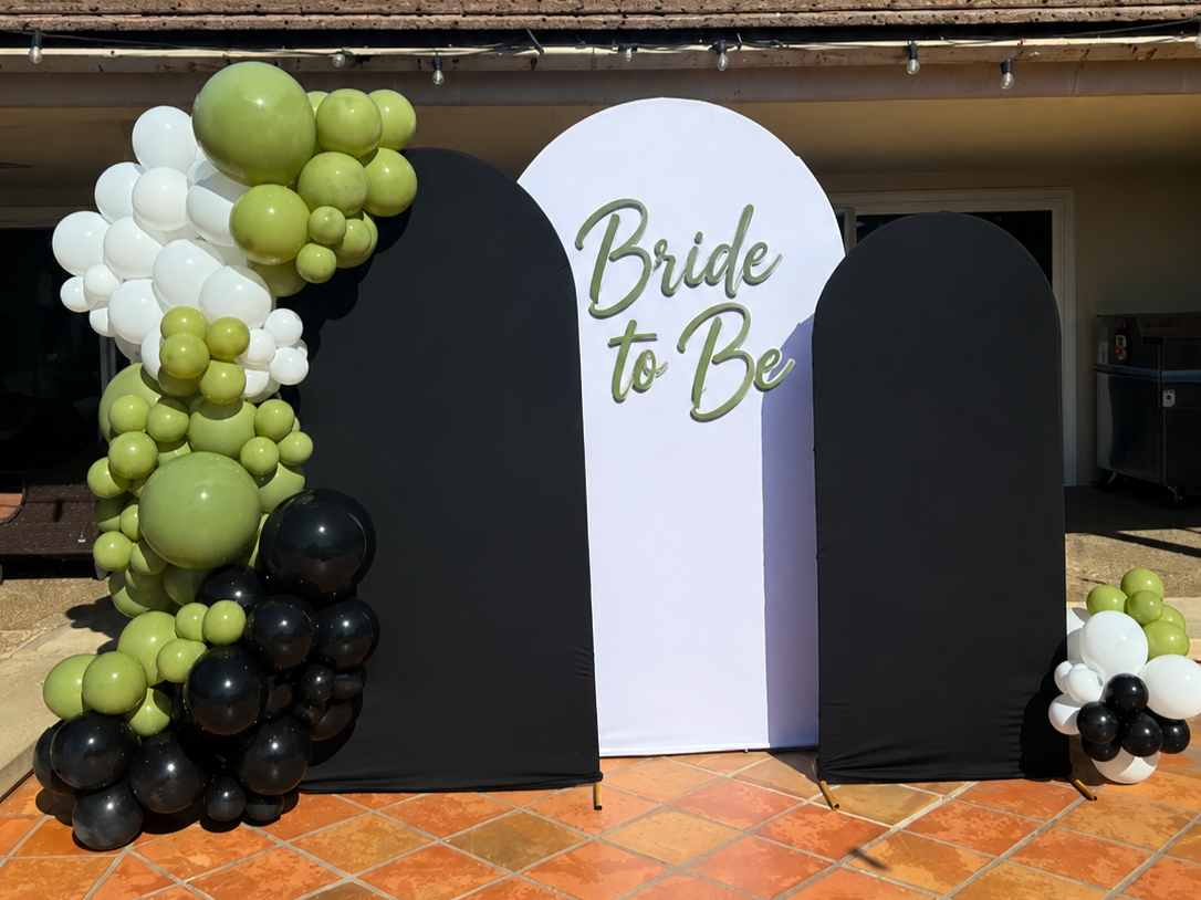 A decorative backdrop for a bridal shower or bachelorette party with a large white arch featuring the words 'Bride to Be' in green lettering. The backdrop is flanked by black panels and decorated with clusters of green, white, and black balloons.