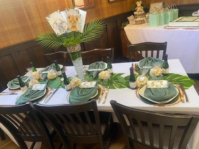 Table decorated with green napkins, gold utensils, and bottles, featuring a tall floral arrangement with drawings of zoo animals on top, in a restaurant or event space with a wood-paneled wall.