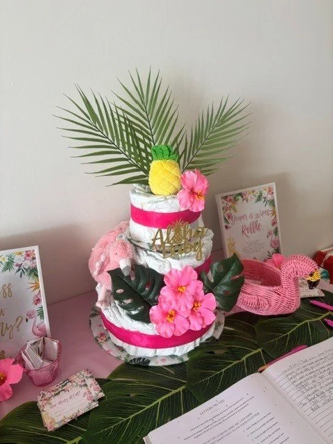 Decorative cake with pink and green tropical theme, topped with a pineapple and palm leaves, surrounded by pink flowers and tropical leaves, with signs and a flamingo figure nearby.