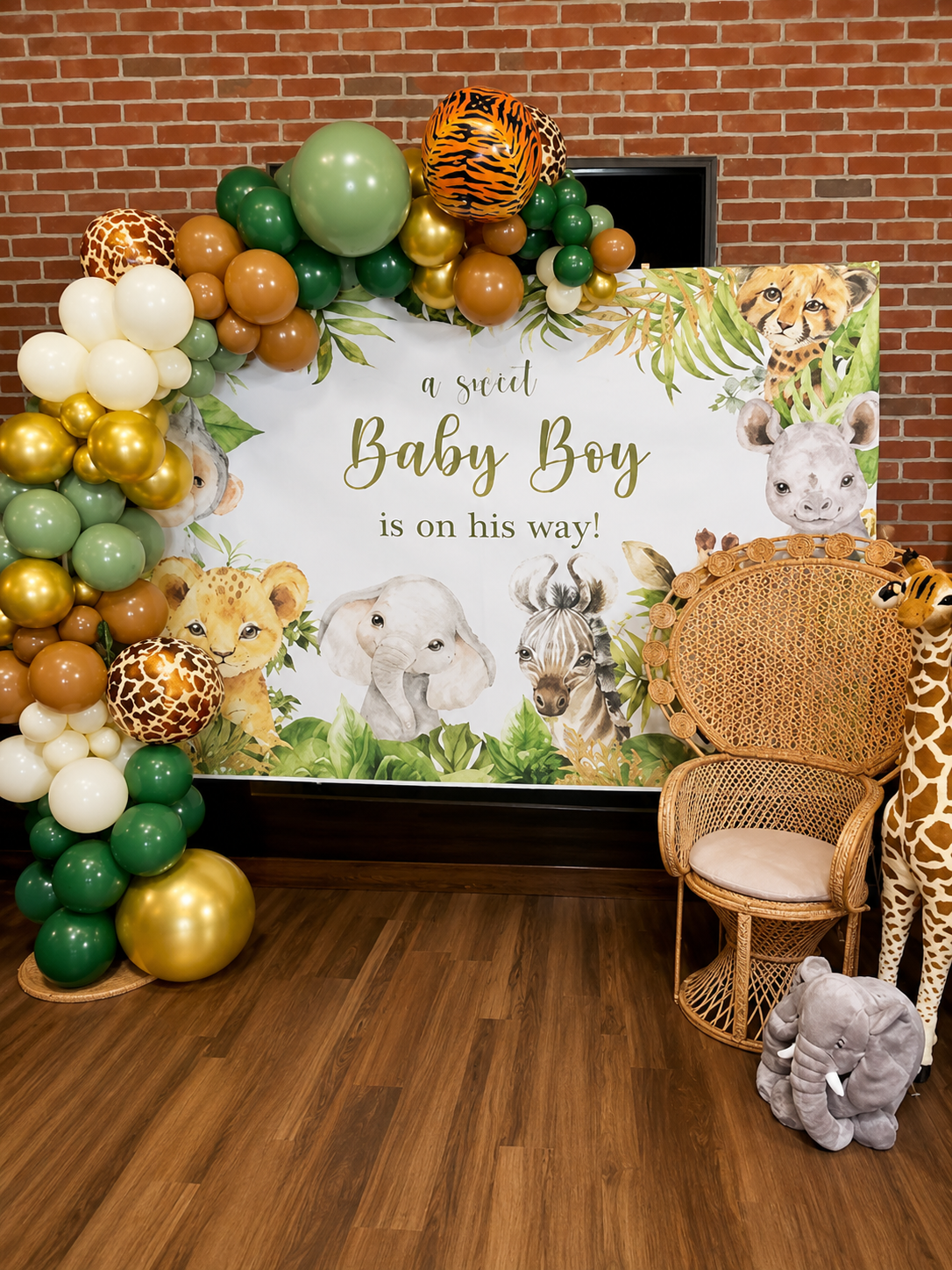 Jungle-themed baby shower backdrop with balloons, stuffed animals, and a wicker chair. The banner reads 'a scrit Baby Boy is on his way!' and features illustrations of a cheetah, giraffe, elephant, zebra, and pig surrounded by green leaves.