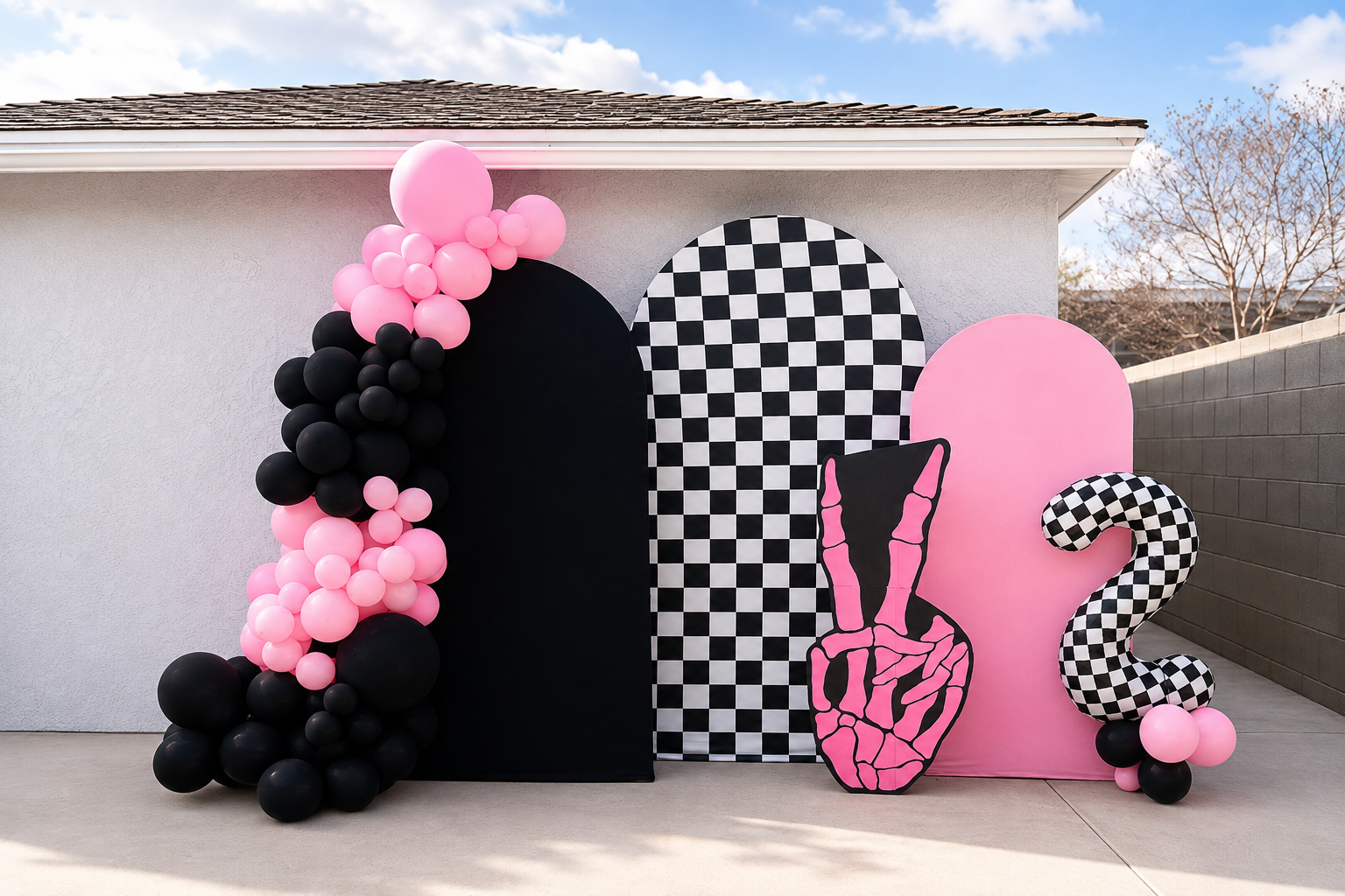 Decorative backdrop with balloons and cutouts, featuring black, pink, and black-and-white checkered patterns, set against the side of a building under a cloudy sky.