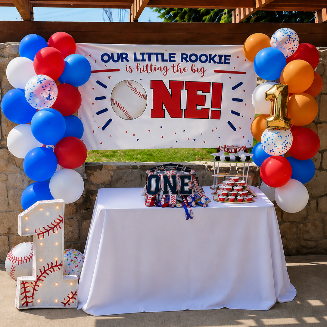 Baseball-themed first birthday party decoration with balloons, a banner reading 'Our little rookie is hitting the big one!', a large illuminated number one, and a table with birthday supplies.