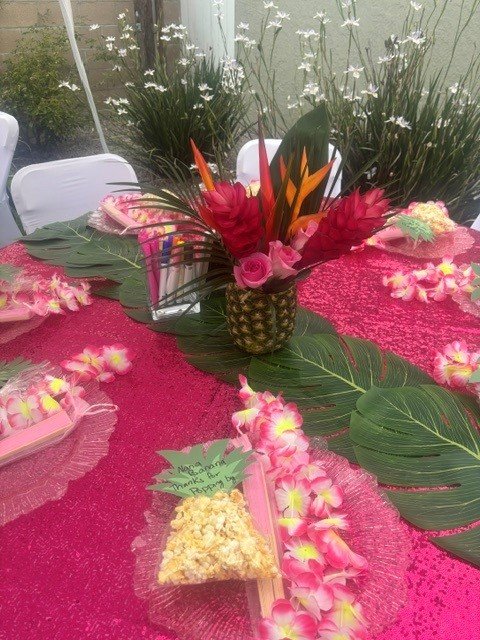 Colorful tropical flower arrangement with red, orange, and pink flowers and green leaves on a pink tablecloth, with small floral decorations and a card.