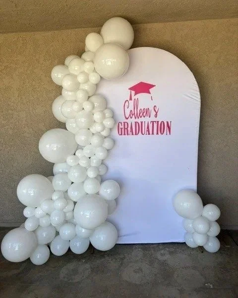 Balloon display with white balloons arranged in clusters beside a white backdrop that has pink text saying 'Colleen's Graduation' with a pink graduation cap graphic.