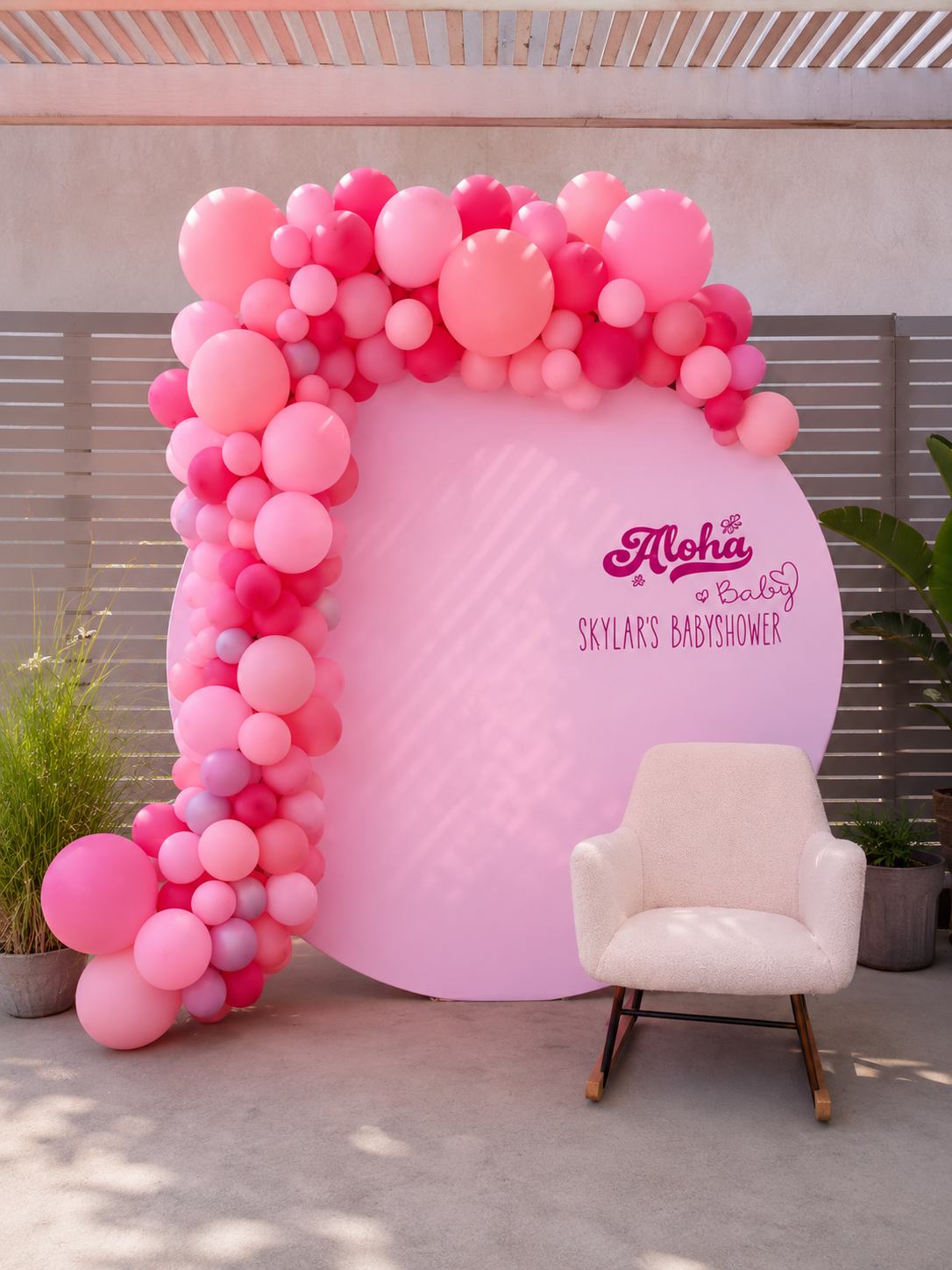 Pink balloon arch and backdrop for a baby shower with the words 'Aloha Baby' and 'Skylar's Babyshower' on the backdrop, and a white armchair nearby.