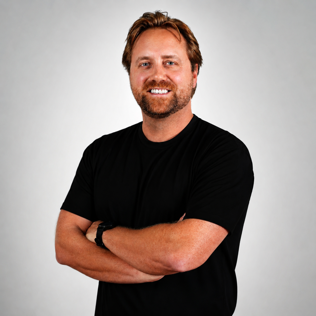 A man with light skin, brown hair, and a beard smiling with arms crossed, wearing a black t-shirt and a watch, standing against a plain light gray background.