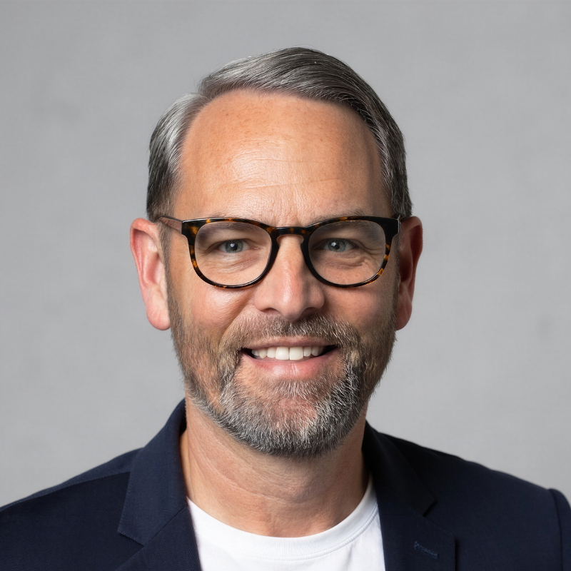 Headshot of a middle-aged man with glasses, short gray hair, and a beard, smiling against a plain gray background.