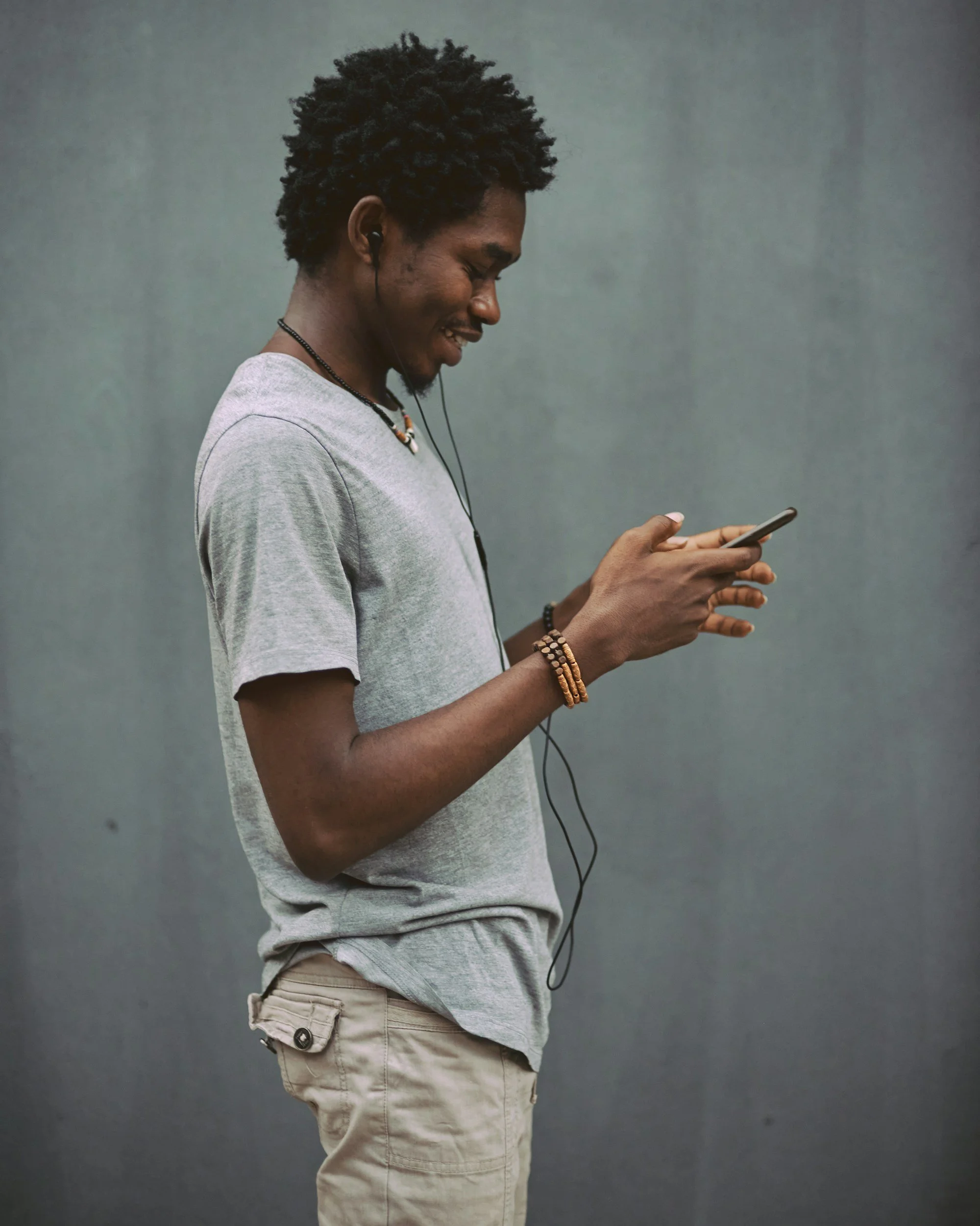 A young man with dark curly hair, wearing a light gray T-shirt, beige cargo shorts, headphones, and beaded jewelry, is looking at his smartphone against a plain gray wall background.
