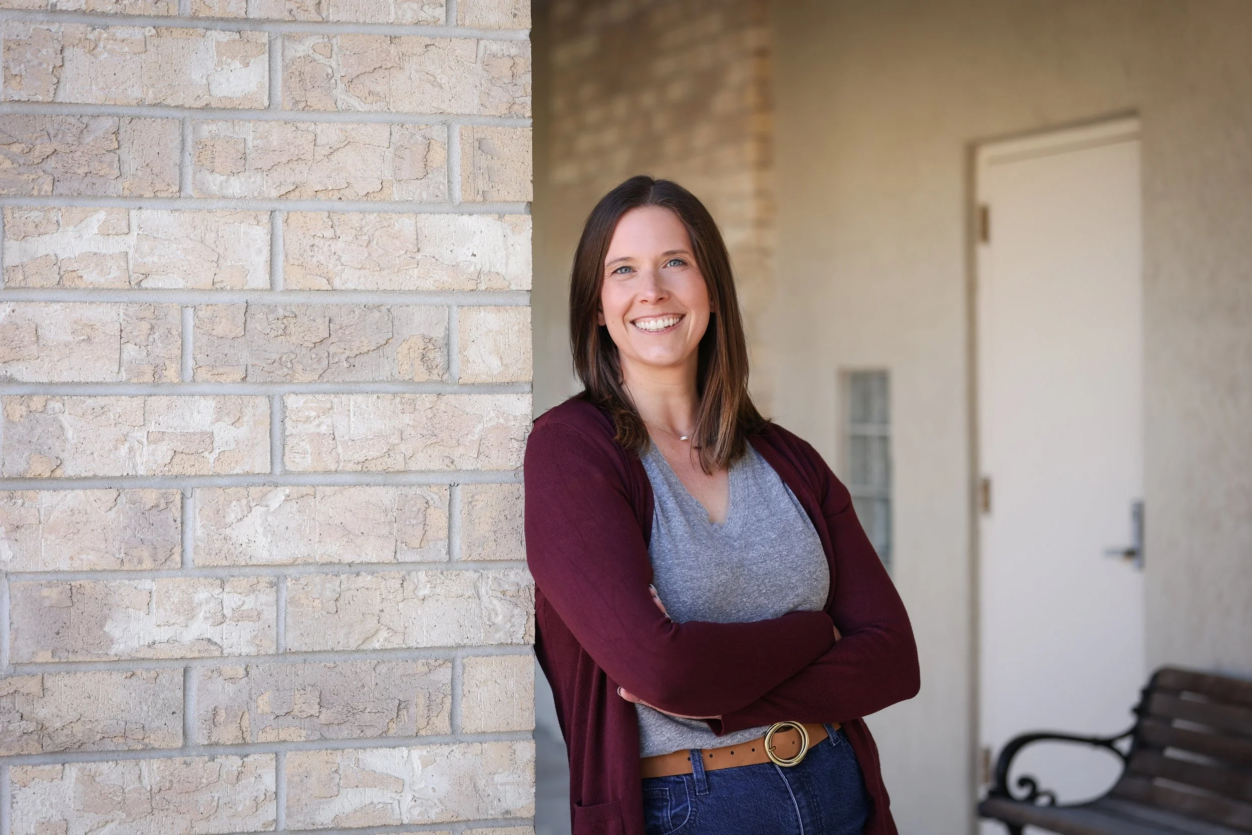 A young woman with shoulder-length brown hair and blue eyes, smiling and standing outdoors next to a beige brick wall. She is wearing a gray t-shirt, a maroon cardigan, and dark blue jeans with a tan belt. Her arms are crossed, and there is a blurred background with a bench and a door.