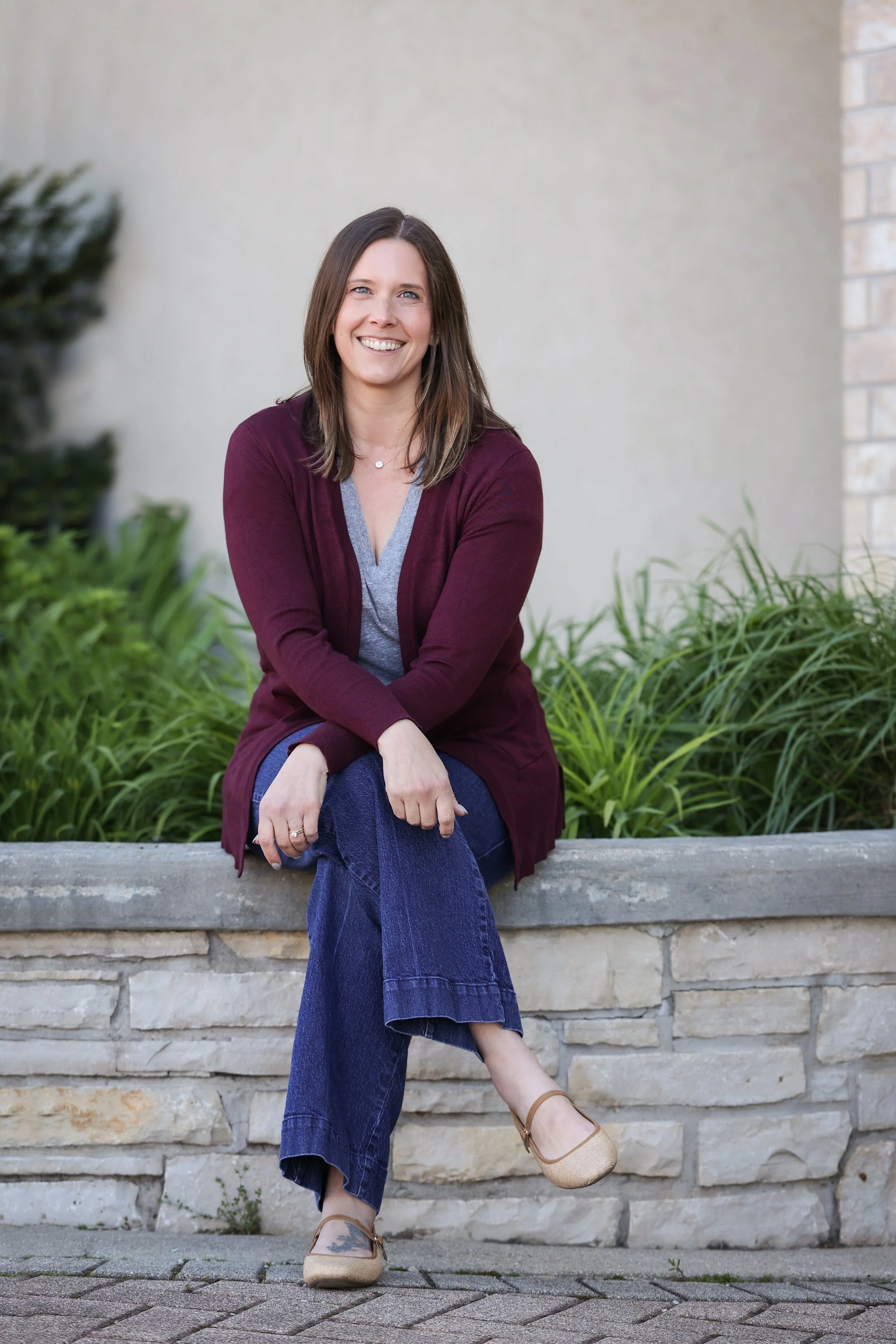 Woman sitting on a stone bench outdoors, smiling, with greenery and a beige wall behind her. She wears a maroon cardigan over a gray top, blue jeans, tan shoes, and has shoulder-length brown hair.