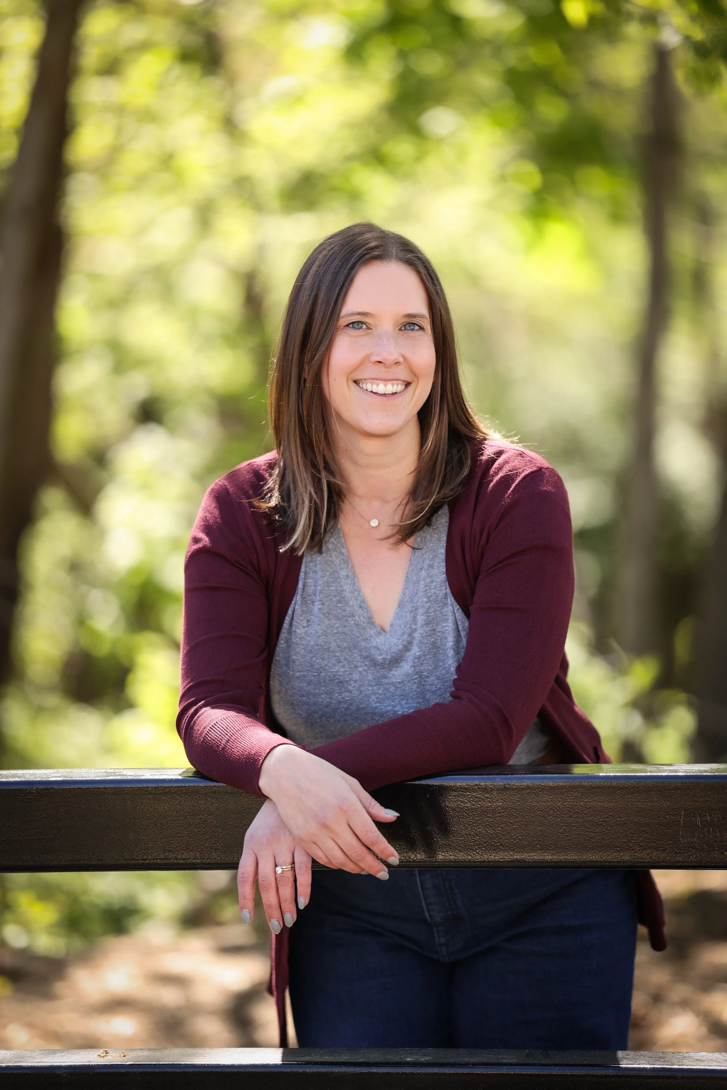 A woman with brown hair and blue eyes smiling outdoors at a park, leaning on a black metal railing, with green trees and sunlight in the background.