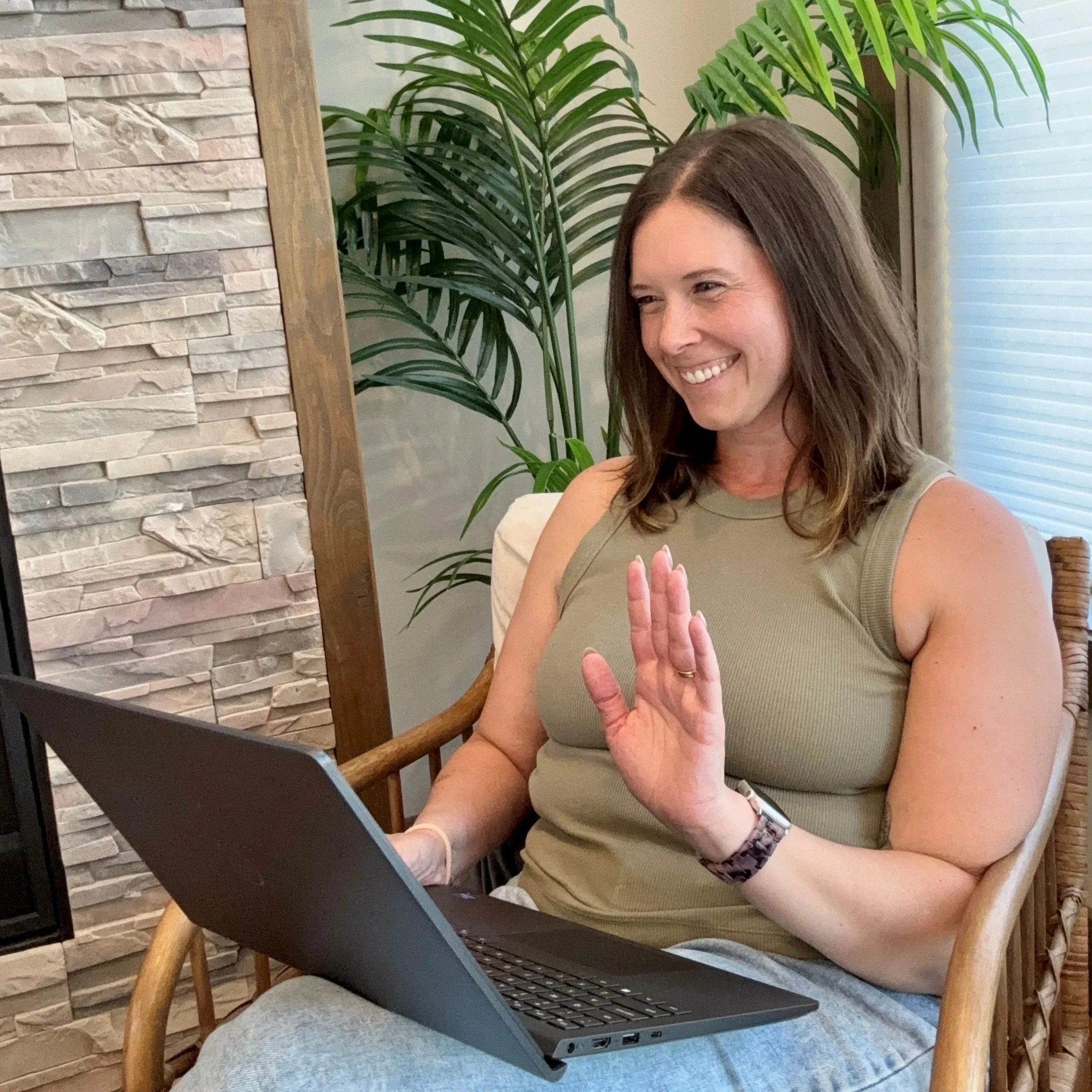 A woman with shoulder-length dark hair smiling and waving while sitting on a brown leather sofa. She is wearing a light-colored shirt over a green top and blue jeans. The background shows a muted green wall with an arched window and some letters partially visible on the wall.