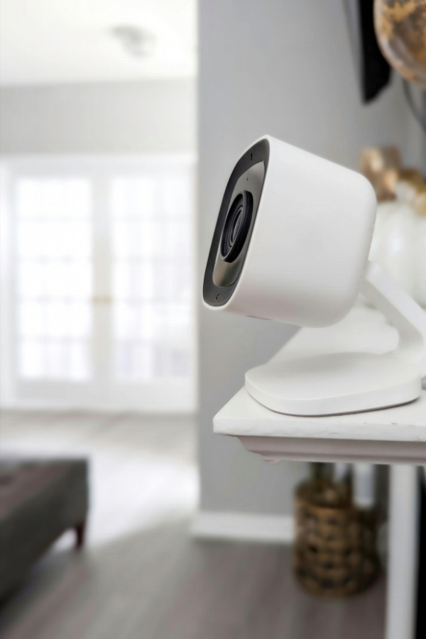 A white security camera mounted on a white shelf in a bright living room with large windows and natural light.