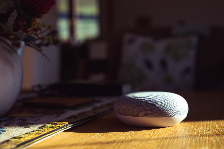 Smart speaker on a wooden table with sunlight shining on it, in a cozy room with a vase of flowers and blurred background.
