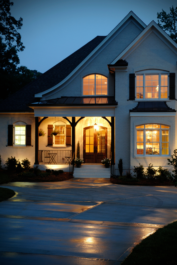 A large two-story house with white brick exterior, lit warmly from inside, with a covered front porch, black shutters, and a curved driveway at dusk.