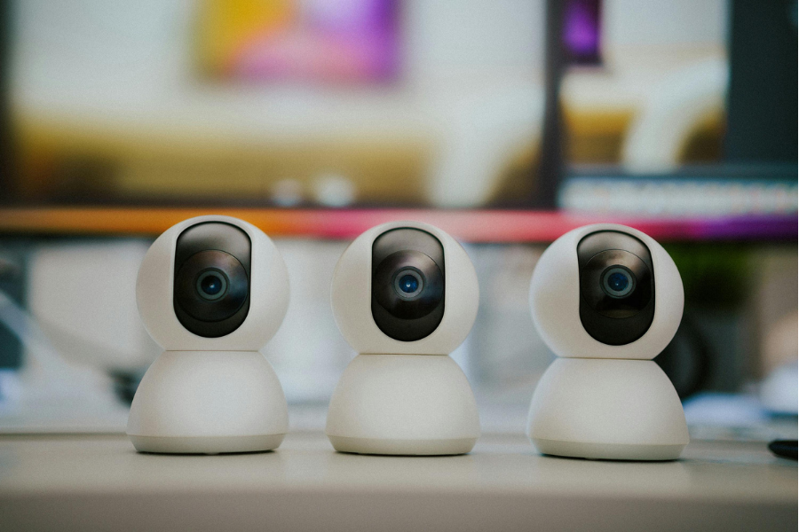 Three white smart security cameras with black lenses placed on a white surface, with a blurred background of an indoor setting.
