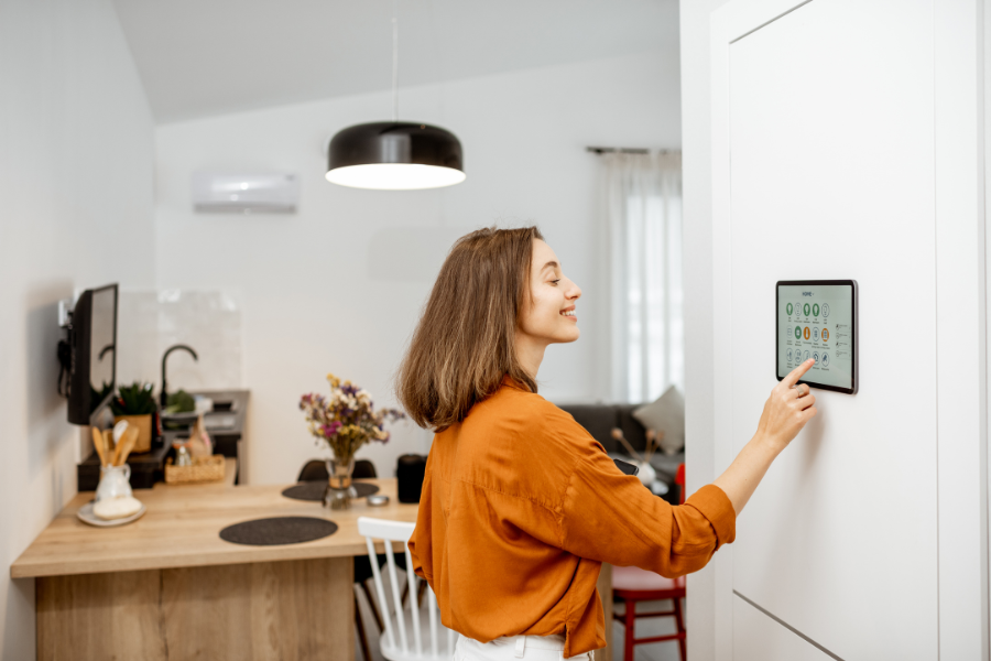 A woman with shoulder-length brown hair in an orange blouse interacts with a touchscreen control panel on a white wall in a modern, bright kitchen.