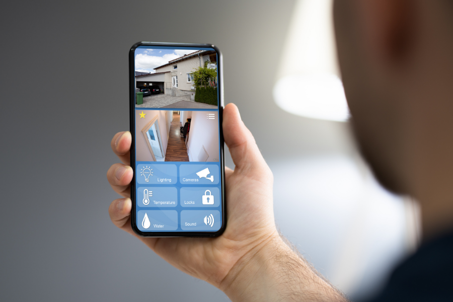 Person holding a smartphone with a home security app open, showing live feeds of the house's exterior and interior.
