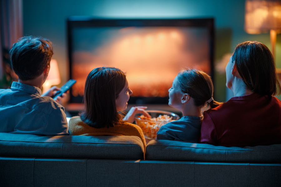 Four people sitting on a sofa watching a movie on a large screen TV in a cozy living room, with a bowl of popcorn in front of them.