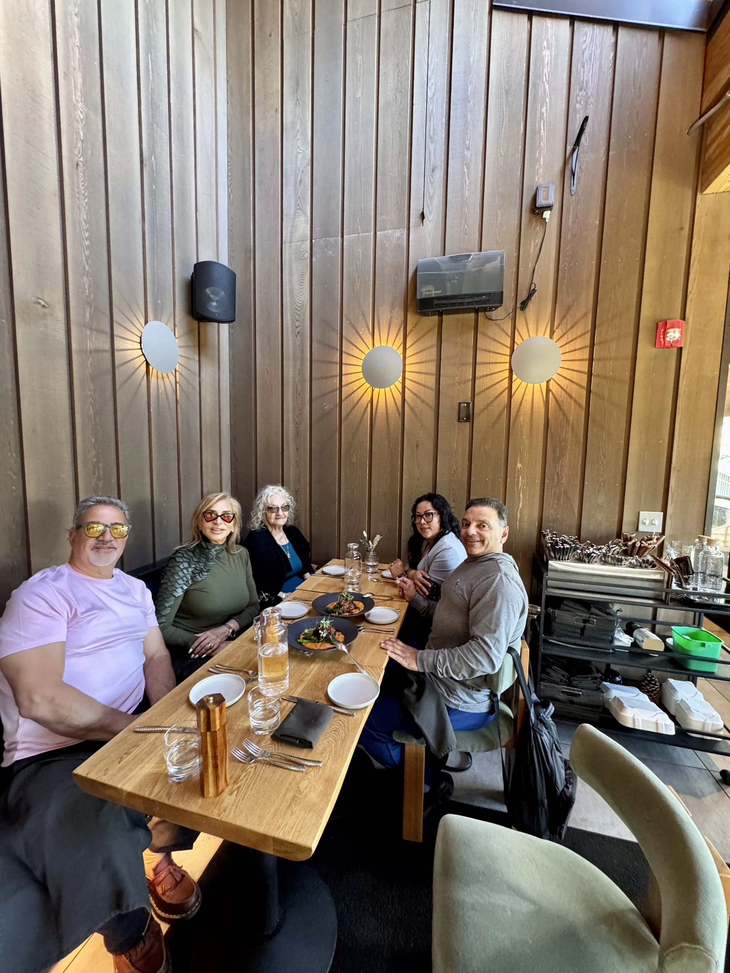 Six people sitting at a long wooden dining table inside a restaurant with wood-paneled walls. They are smiling and have plates of food in front of them, with some water bottles on the table.